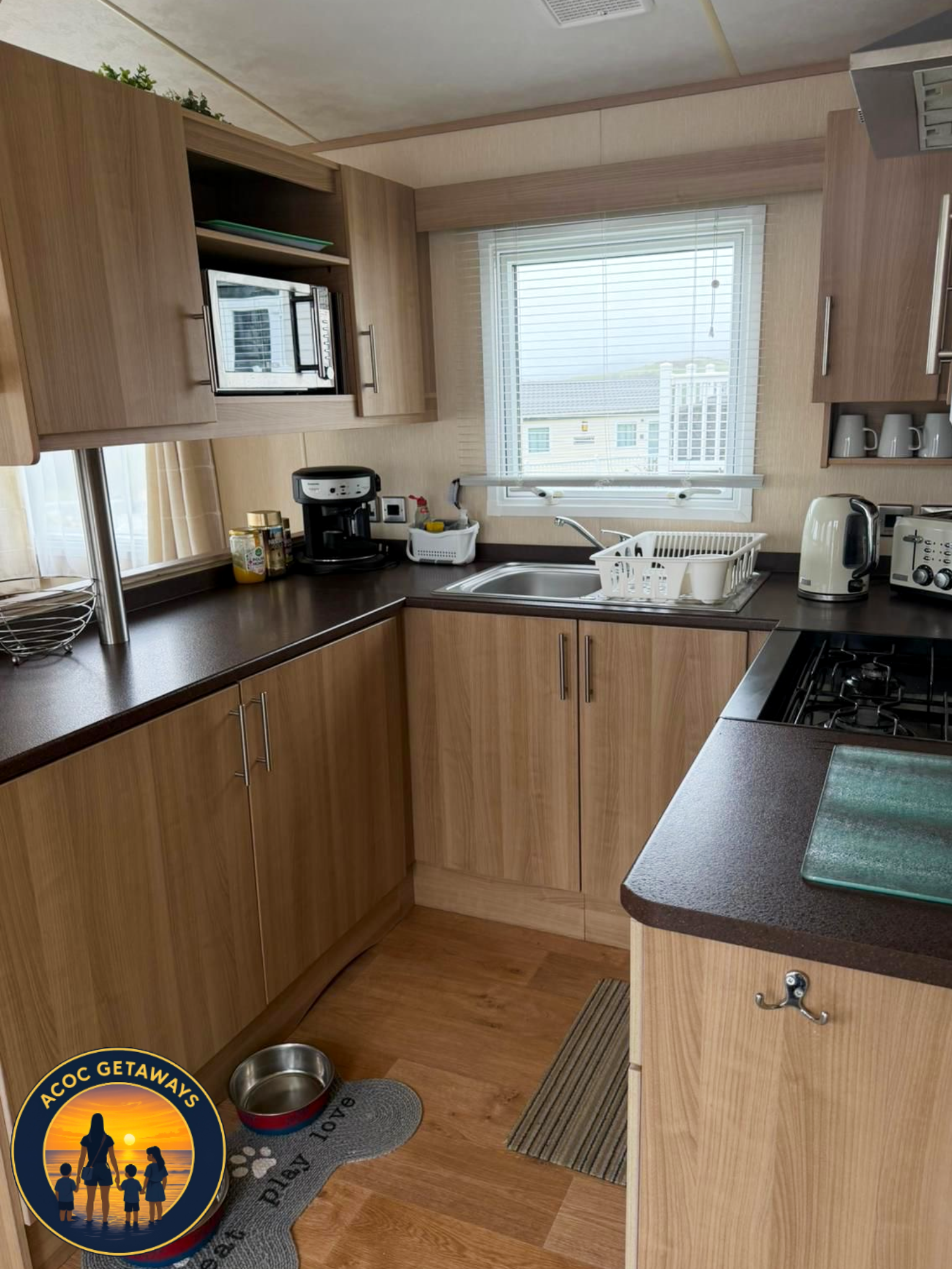 Kitchen with wooden cabinets, a window above the sink, and various appliances including a microwave, coffee maker, toaster, and kettle. A pet food bowl sits on a mat on the floor near the cabinets.