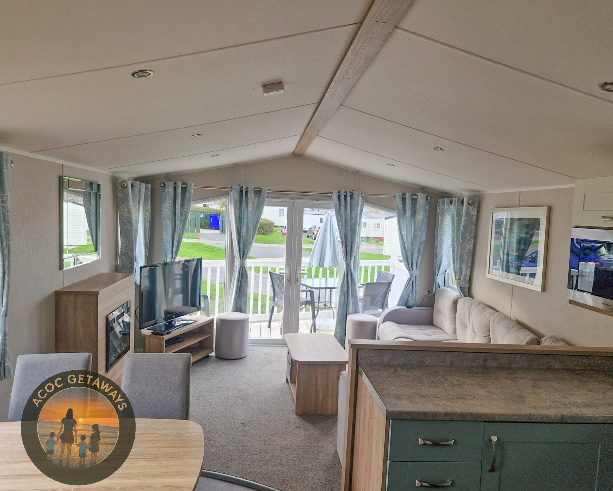 Living room with large windows and sliding glass door leading to a balcony with outdoor furniture. Inside, there is a beige sectional sofa, a flat-screen TV on a wooden stand, a fireplace, and a round ottoman. Light blue patterned curtains frame the windows, and a wood-beamed ceiling overhead.