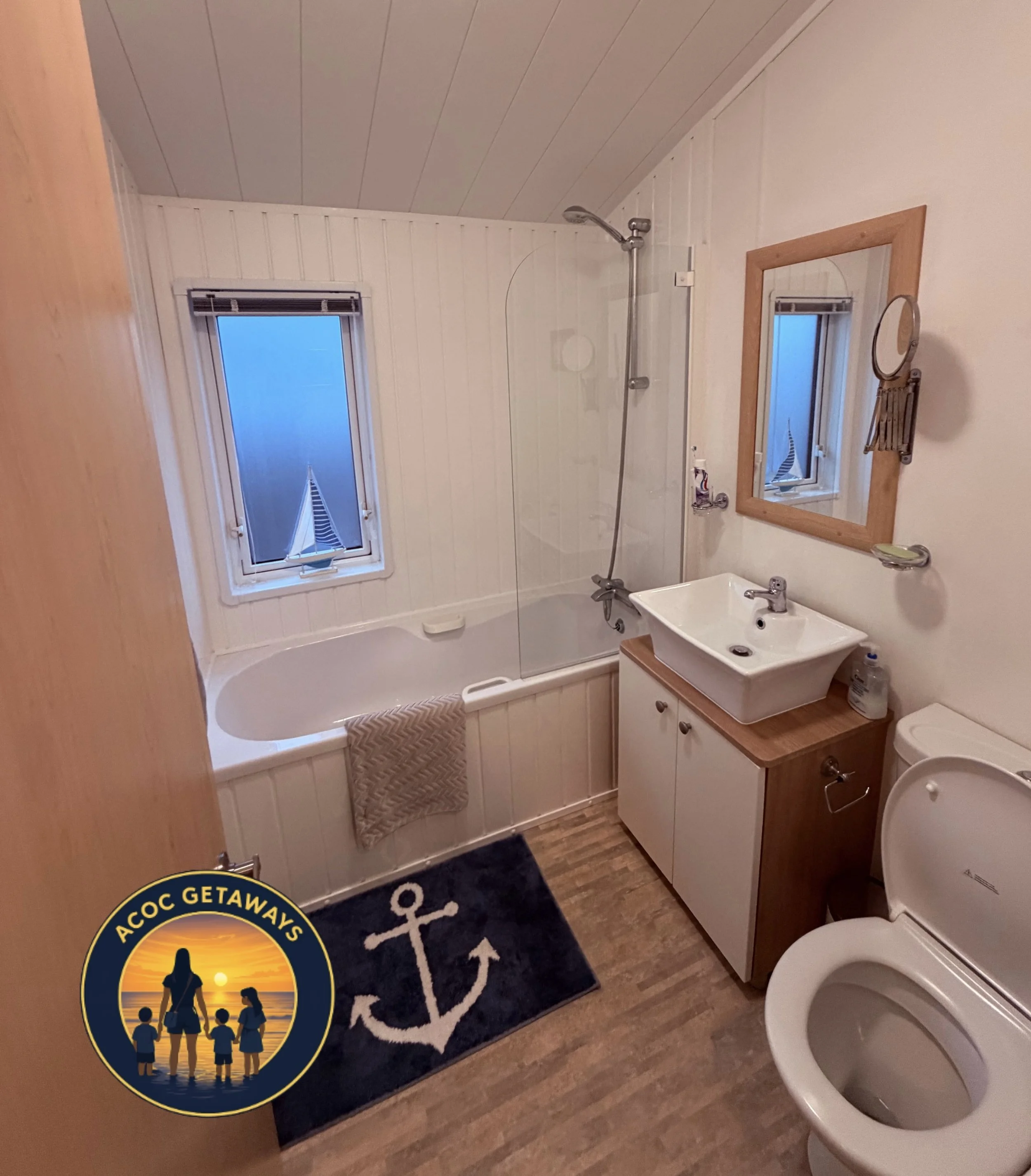 A small nautical-themed bathroom with a bathtub, shower, sink, mirror, and toilet. The rug features an anchor design, and the wall art includes a round mirror on a wooden frame and a smaller mirror hanging beside it.