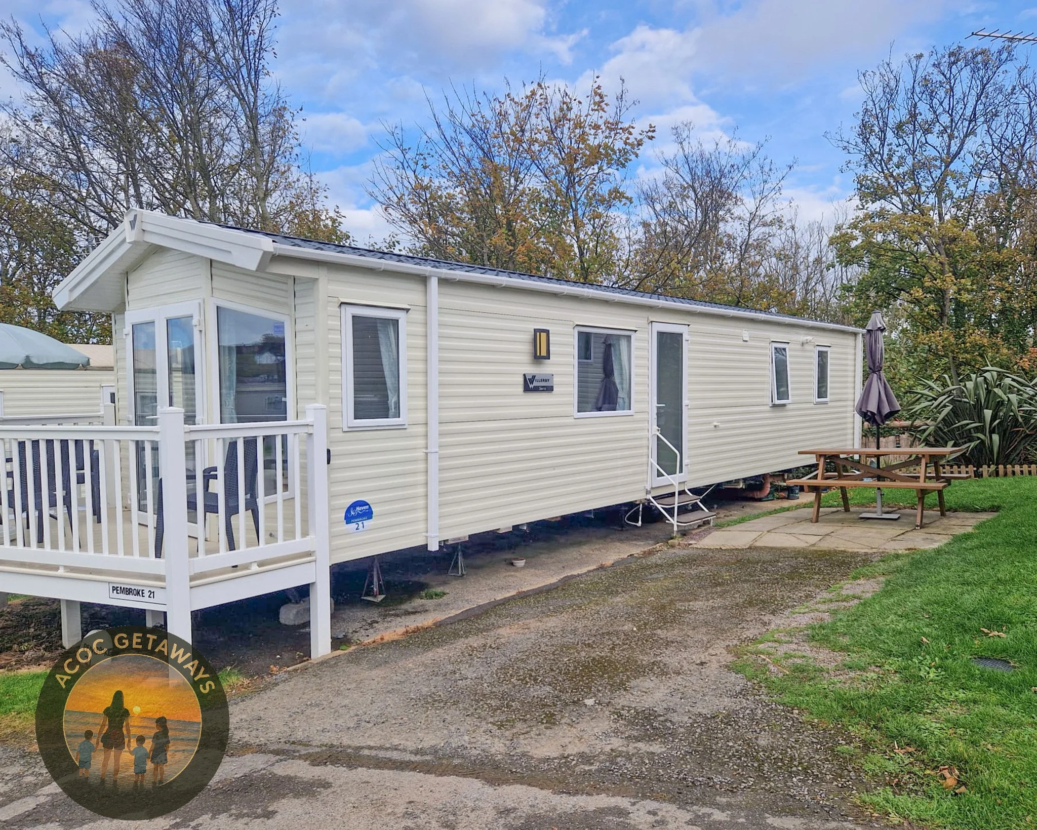 A white mobile home with a small porch, several windows, and an outdoor seating area with a table and umbrella, set in a grassy area with trees in the background on a cloudy day.
