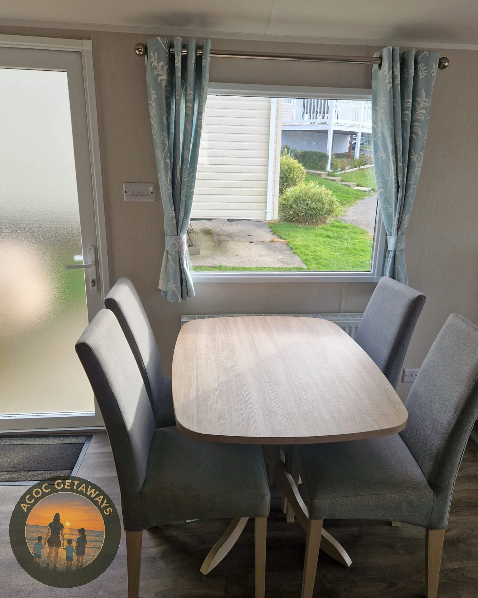 A wooden dining table with four gray upholstered chairs inside a room, with a window and a door leading outside. The window has blue and white curtains, and there is greenery and a pathway outside.
