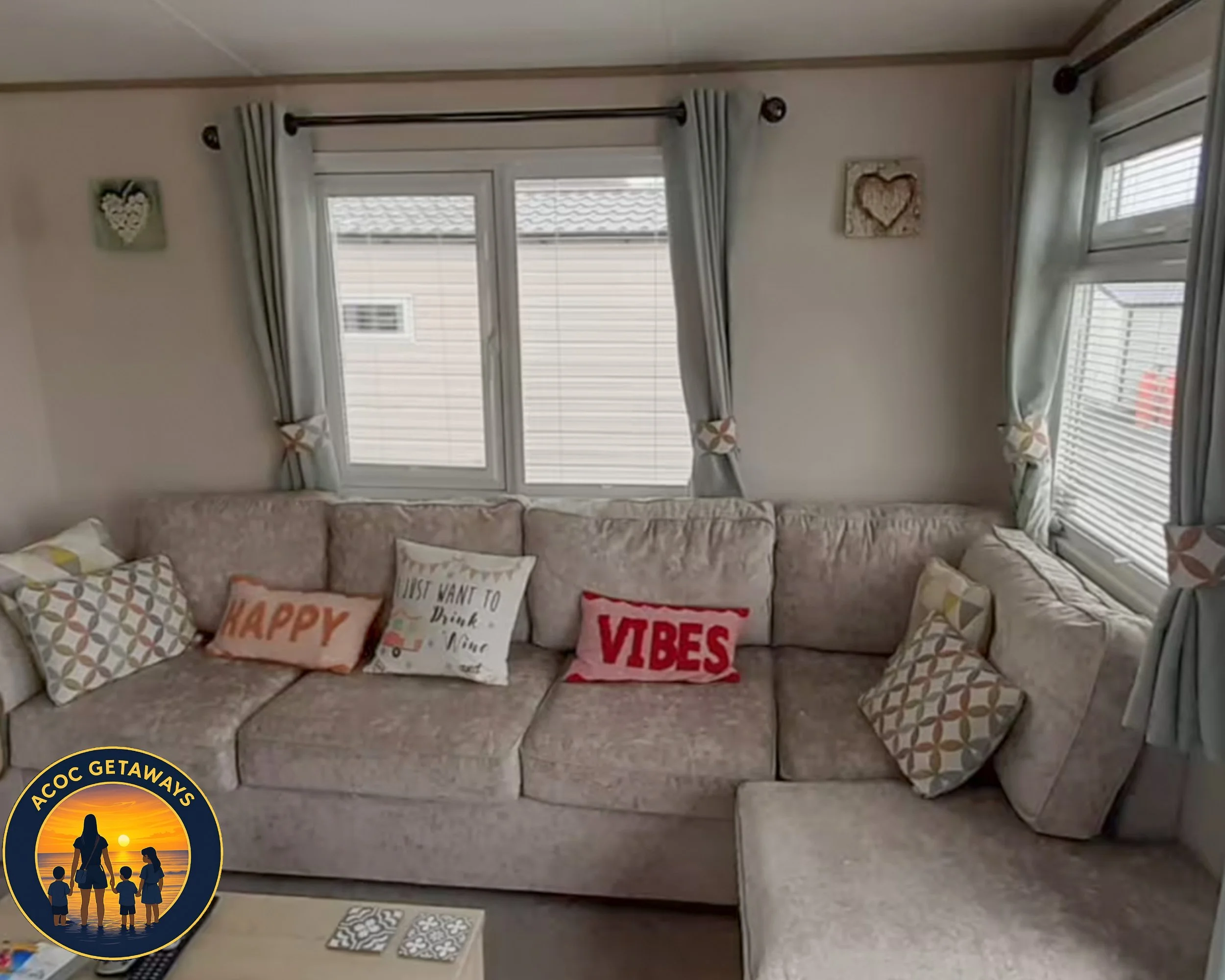 Living room with a beige L-shaped sectional sofa, decorated with throw pillows that say 'HAPPY', 'VIBES', and 'Just want to think, wine'. There are blue curtains with tiebacks over windows, and wall art with hearts. Part of a beige coffee table with 