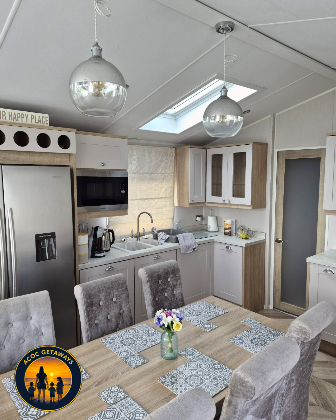 Kitchen with a dining table and six upholstered chairs, a rustic wooden table with decorative placemats and a flower vase, white cabinets with glass doors, a microwave, a refrigerator, a sink, and skylight providing natural light.