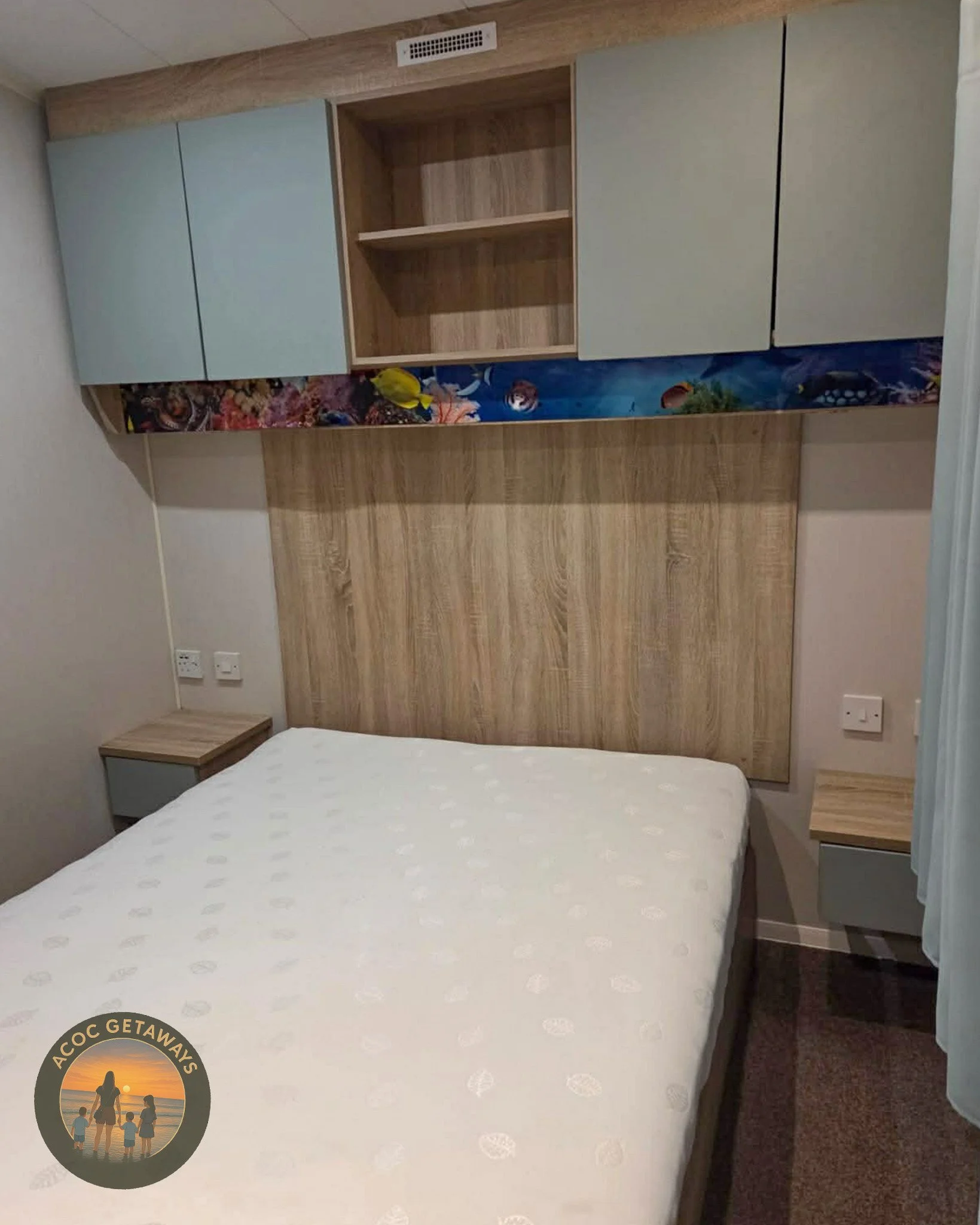 A bedroom with a wooden headboard, two small tucked bedside tables, and a wall-mounted storage unit with closed cabinets, open shelves, and an underwater-themed decorative panel. The bed has a white mattress, and there are electrical outlets on either side of the bed.