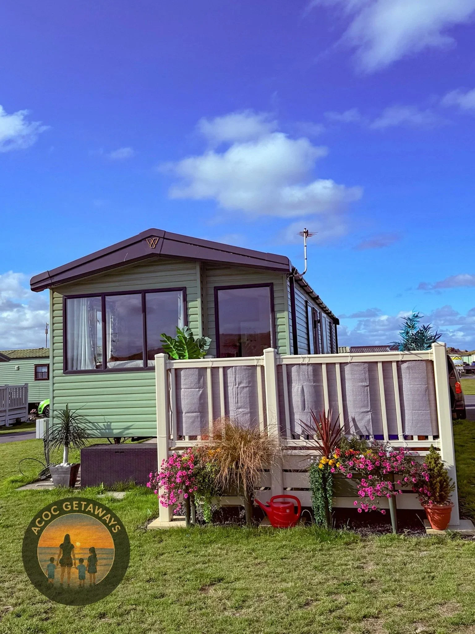 A small light green mobile home with large windows, surrounded by a fenced deck adorned with potted plants and flowers, set on a grassy area under a bright blue sky with scattered clouds.