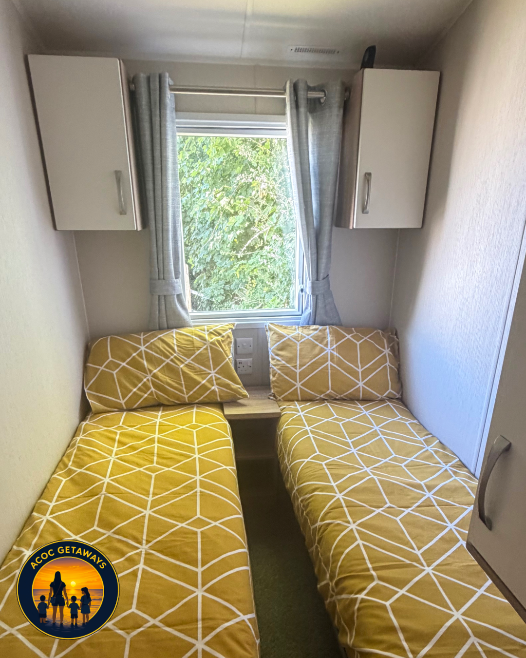 Small bedroom with two single beds with yellow geometric-patterned bedding and matching pillows, centered by a wooden nightstand, a window with grey curtains and green foliage outside, and overhead cabinets.