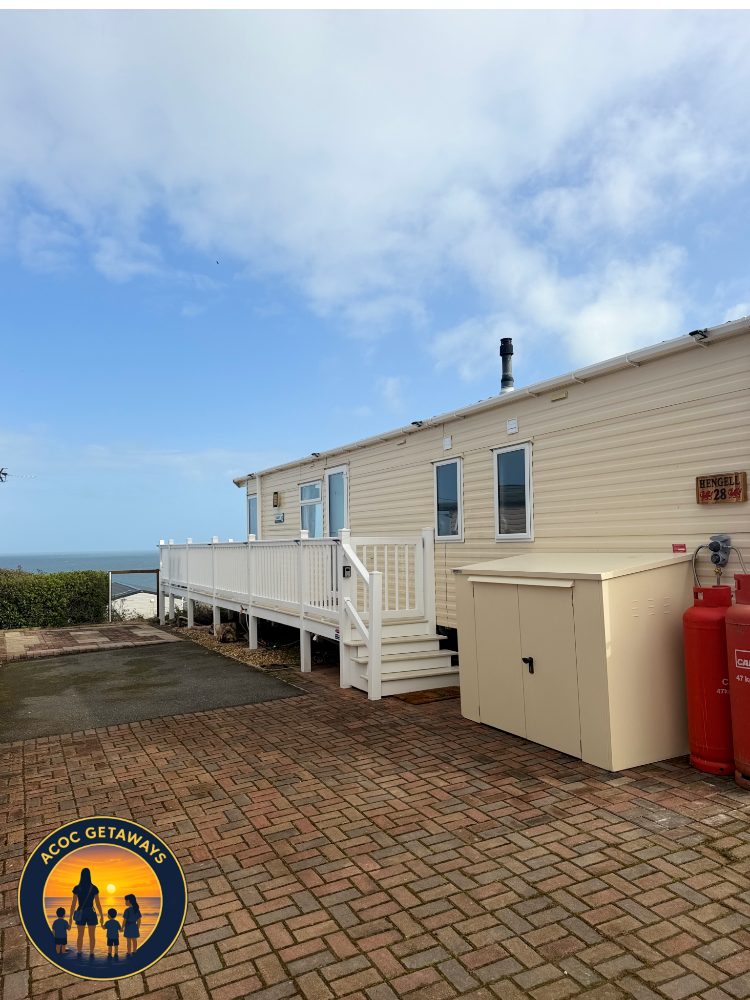 View of the side of a house with a porch, three windows, a chimney, and a sign, with a cloudy sky in the background. The image is partially covered by a round emblem in the bottom left corner featuring a silhouette of a woman with three children at s