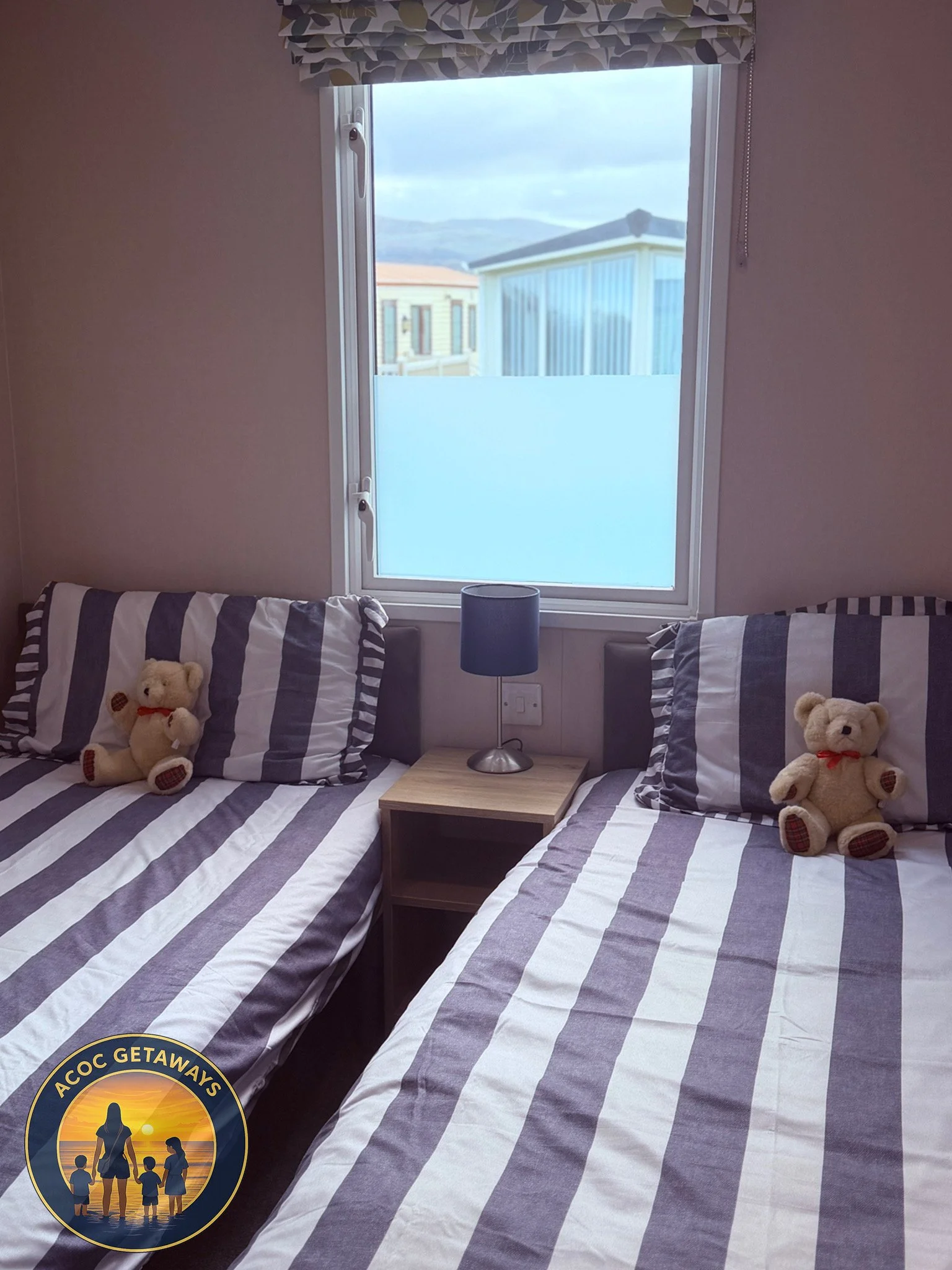 Two single beds with striped bedding and teddy bears, separated by a small bedside table with a lamp, in front of a window showing neighboring buildings and mountains in the distance.