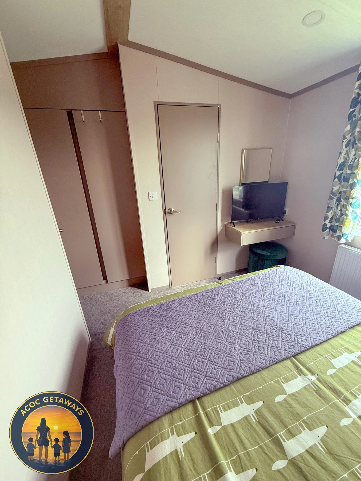 A bedroom with a bed covered in a purple quilt and a green bedspread with white animal silhouettes. A small black flat-screen TV is on a wall-mounted shelf, with a green ottoman below. A window with patterned curtains is on the right, and an open clo