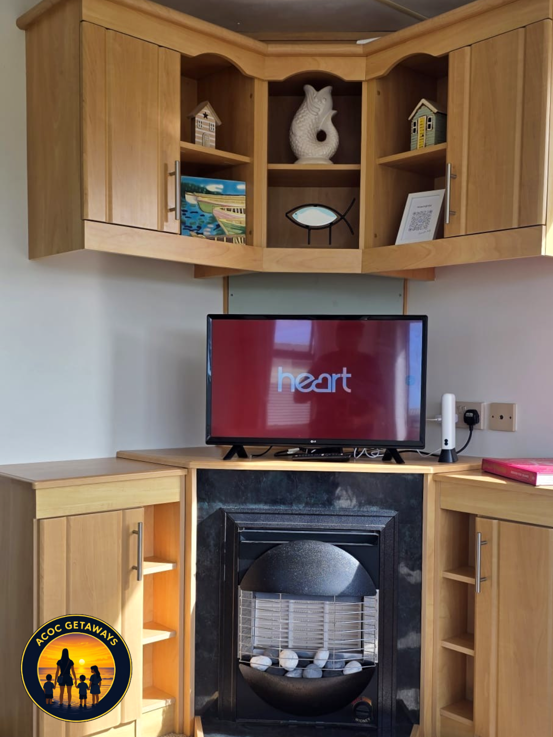 A living room fireplace with a mounted wooden cabinet above it. The cabinet contains decorative items including a ceramic fish, a small house model, a framed QR code, and a painting. The fireplace has a black surround with white stones inside, and a 