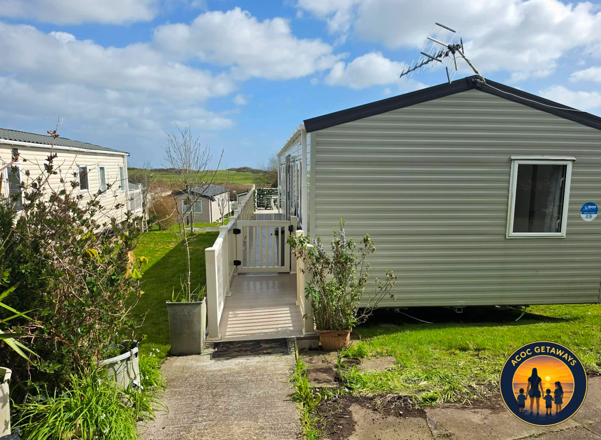 View of a backyard with a garden, a small deck, and multiple mobile homes with antennas on the roofs. The sky is blue with some clouds.