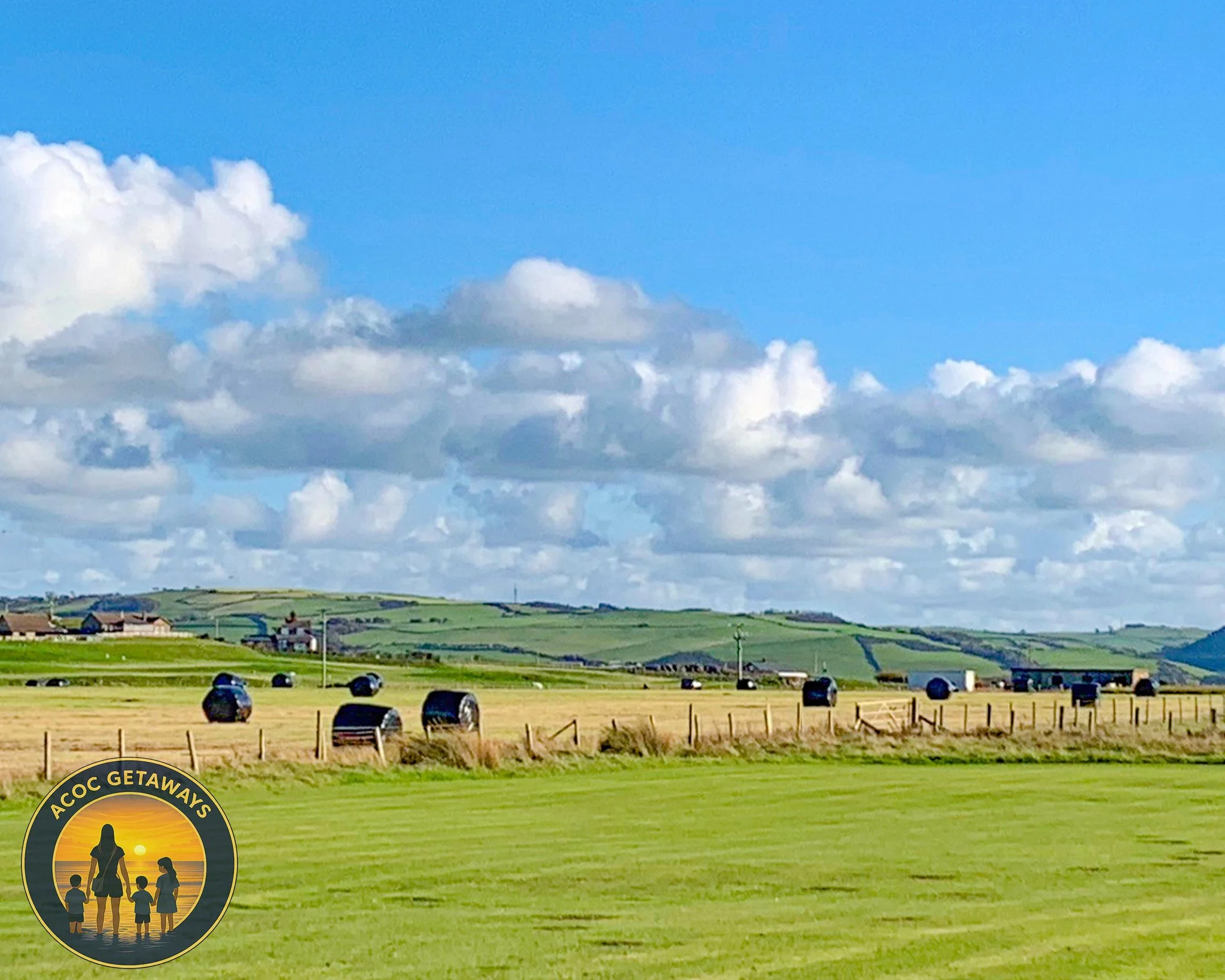 A scenic landscape with rolling green hills under a partly cloudy sky, with patches of farmland and hay bales in the field, and a logo in the bottom left corner showing silhouettes of a person with children during sunset.