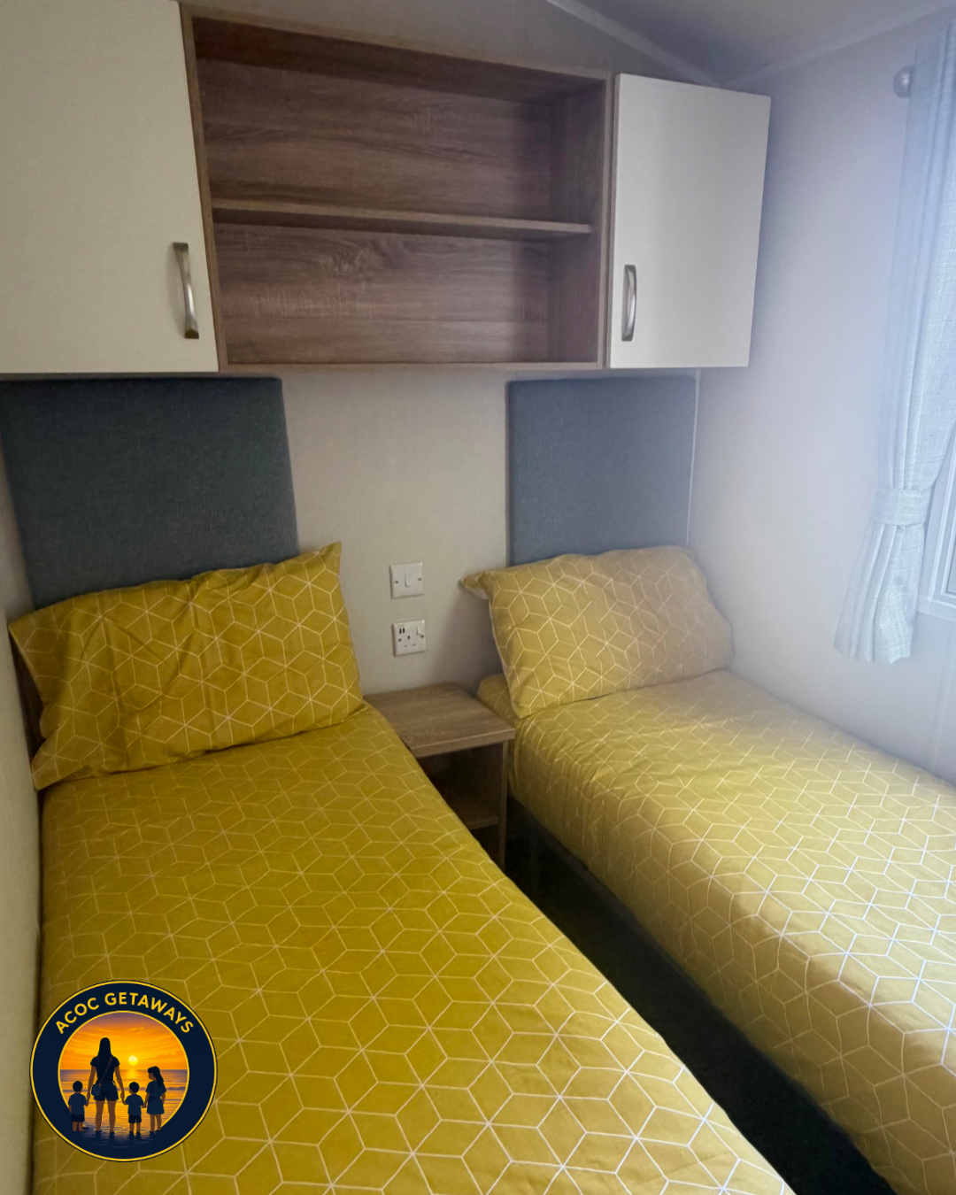 Two single beds with yellow geometric bedspreads and pillows, a small nightstand, built-in wooden and white wall cabinets, and a window with white curtains in a bedroom.