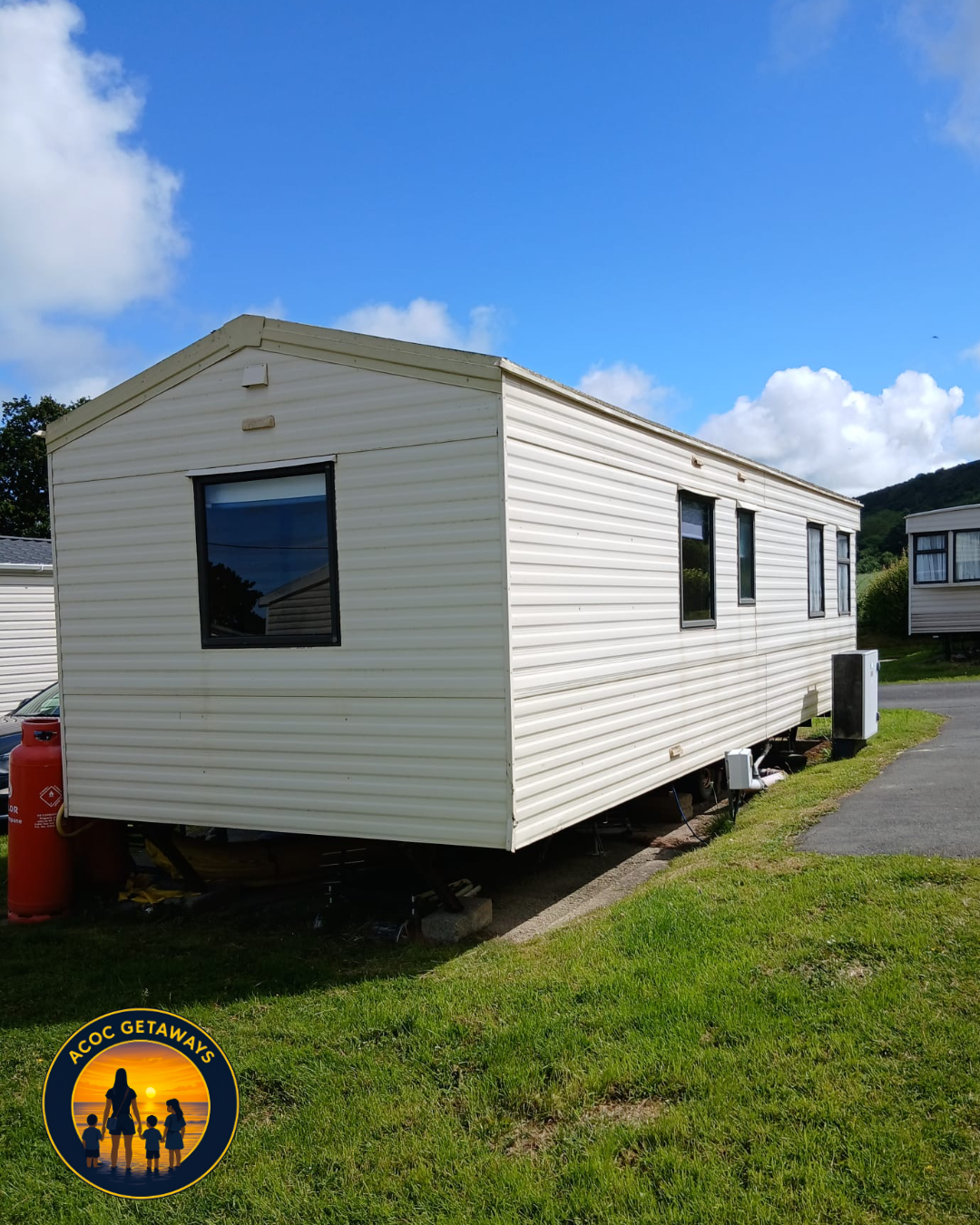 A white mobile home with black window frames on a grassy lot under a blue sky with clouds.