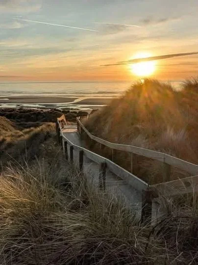 A scenic view of sand dunes with a wooden pathway leading towards the ocean during sunset.