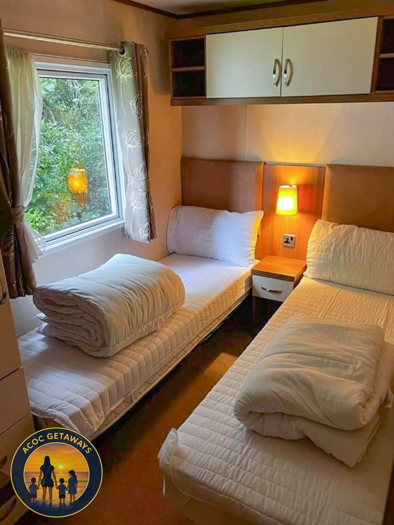 A cozy bedroom with two single beds, each with a pillow and a rolled-up duvet. A window shows greenery outside, and a small nightstand with a lamp is between the beds. There are built-in shelves and cabinets above the beds.