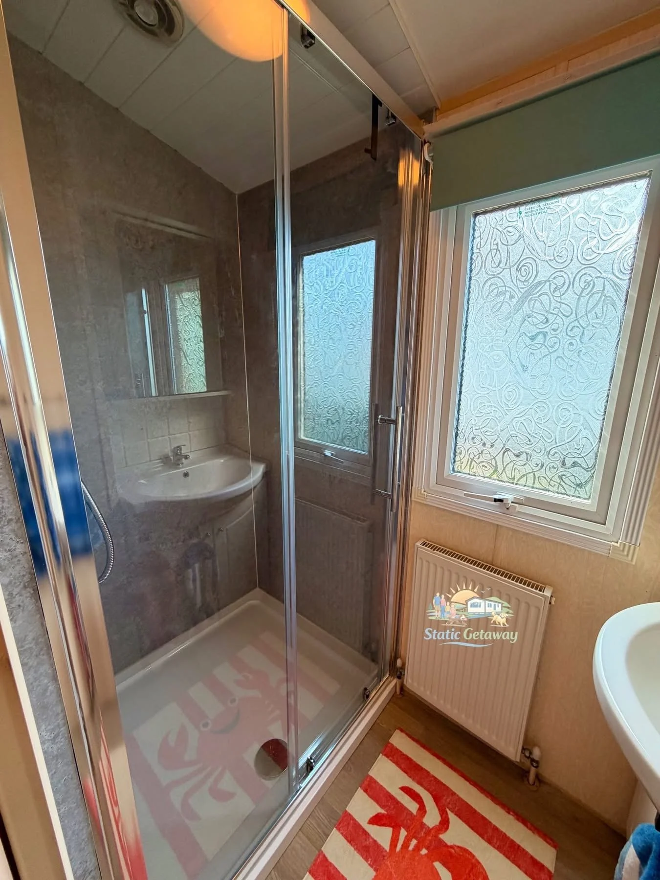 Small bathroom with a shower stall with glass doors, a window with patterned frosted glass, and a radiator below the window. There is a red and white striped rug with a crab design on the bathroom floor.