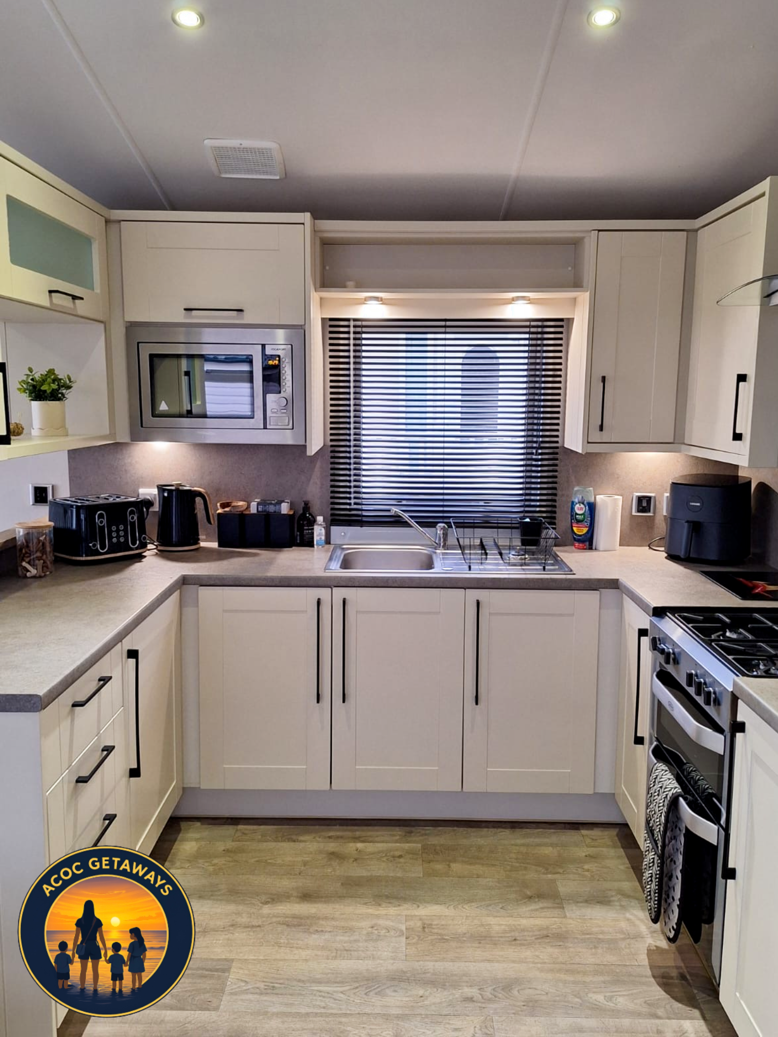 Modern kitchen with white cabinets, black handles, a stainless steel microwave, black toaster, electric kettle, sink under a window with black blinds, stove, and various small appliances and items on the countertops.