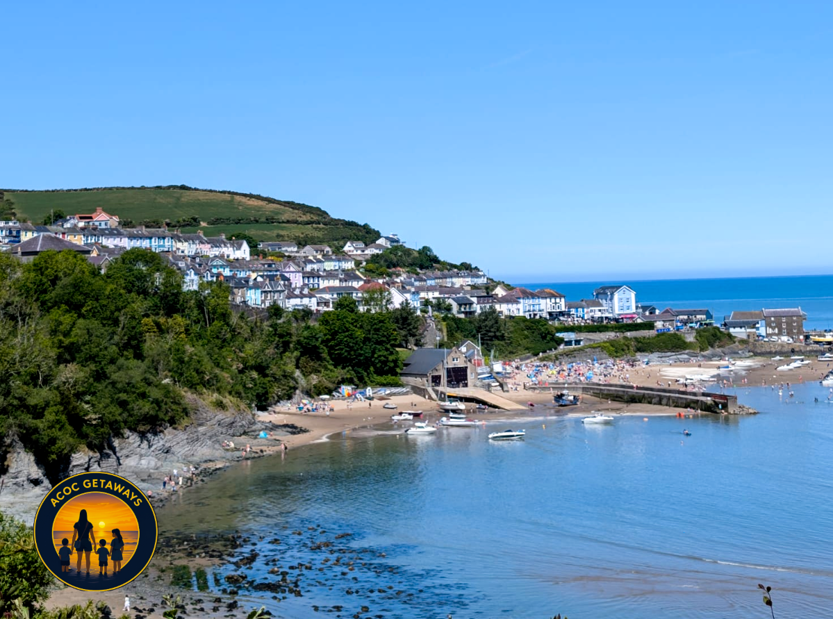 A coastal town with colorful houses on a hillside overlooking a sandy beach and calm water, with boats and people enjoying a sunny day.