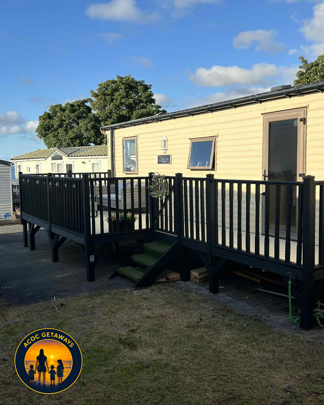 A yellow mobile home with a black deck and stairs, decorated with a wreath, situated outdoors under a blue sky with some clouds. There is a grassy area in front and other similar mobile homes in the background.