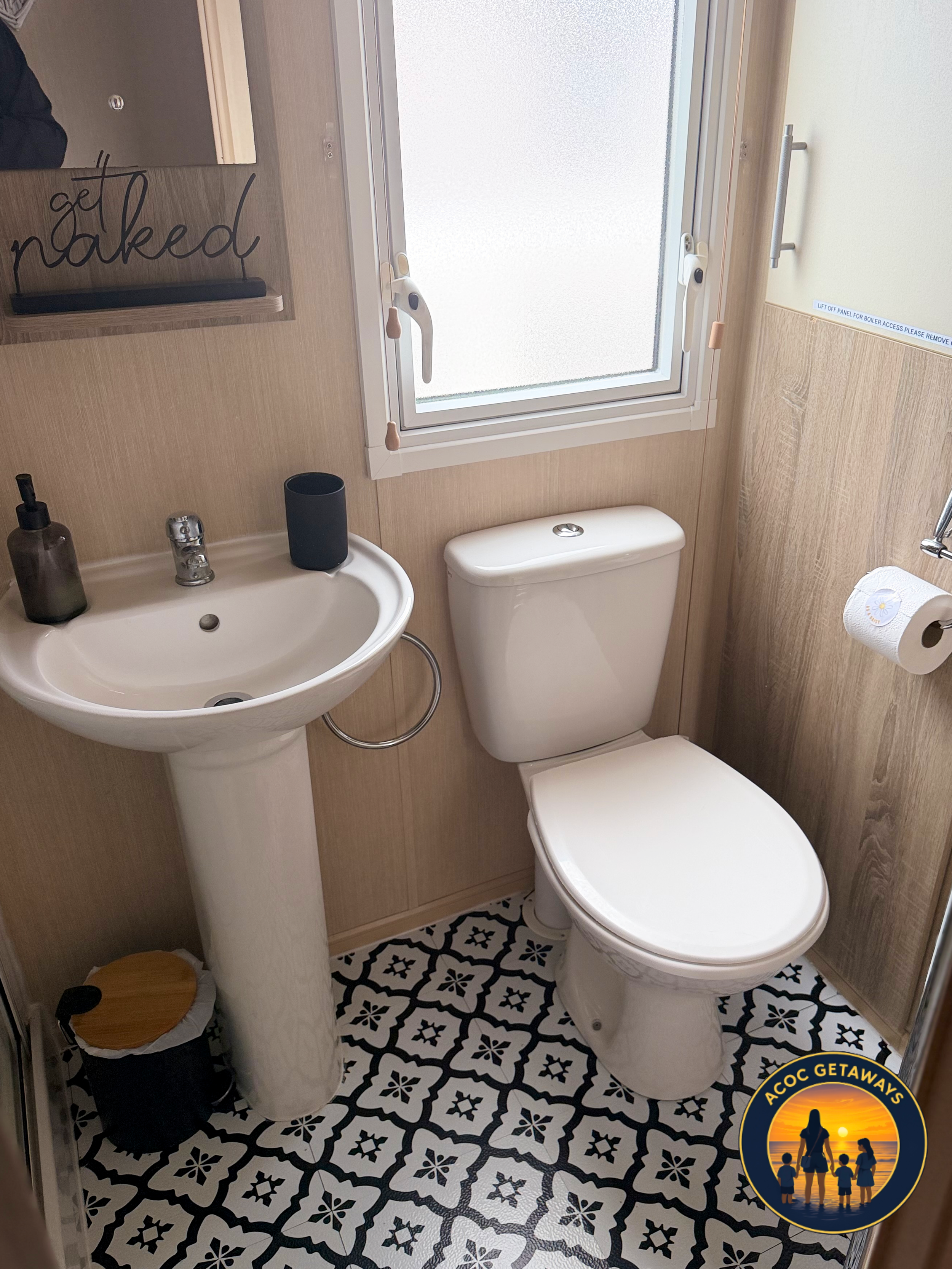 A small bathroom with a white toilet, a white pedestal sink, and a patterned black and white floor. A window with frosted glass above the toilet. A mirror above the sink with a sign that says 'get naked' on it. A soap dispenser and a black cup on the