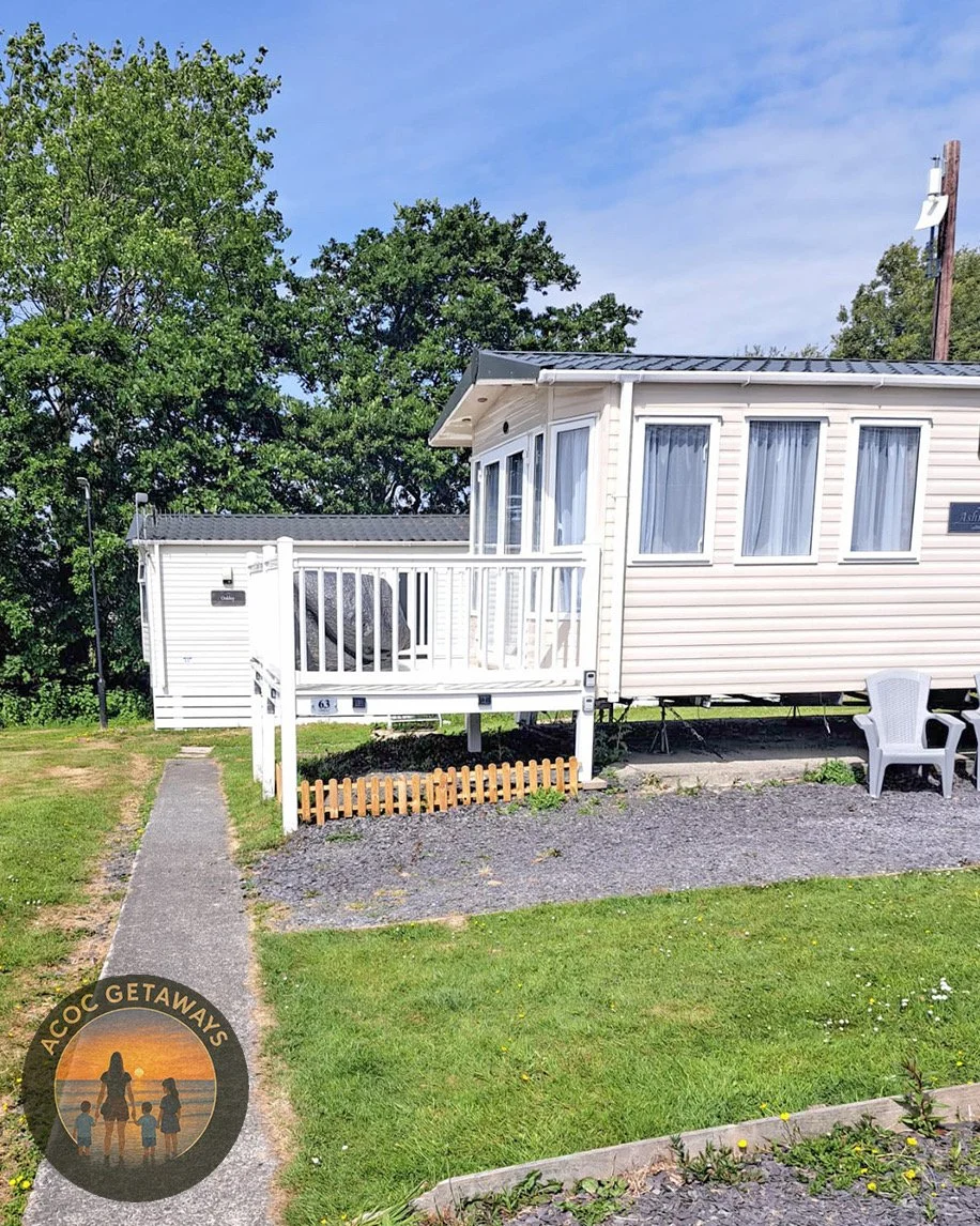 A white mobile home with multiple windows, a small front porch with a white railing, and an outdoor plastic chair. There is a gravel area with a small wooden fence and a sidewalk leading to the home. Green grass, trees, and a blue sky are in the background.