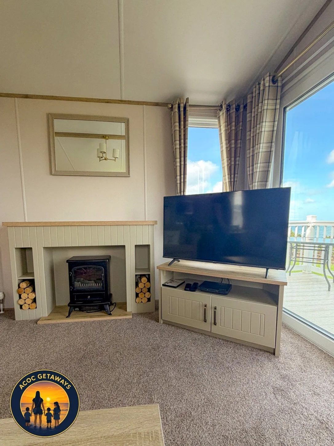 Living room with a fireplace, a large flat-screen TV on a stand, and a sliding glass door leading to a balcony with outdoor furniture. The room has beige walls, checked curtains, and a mirror above the fireplace.