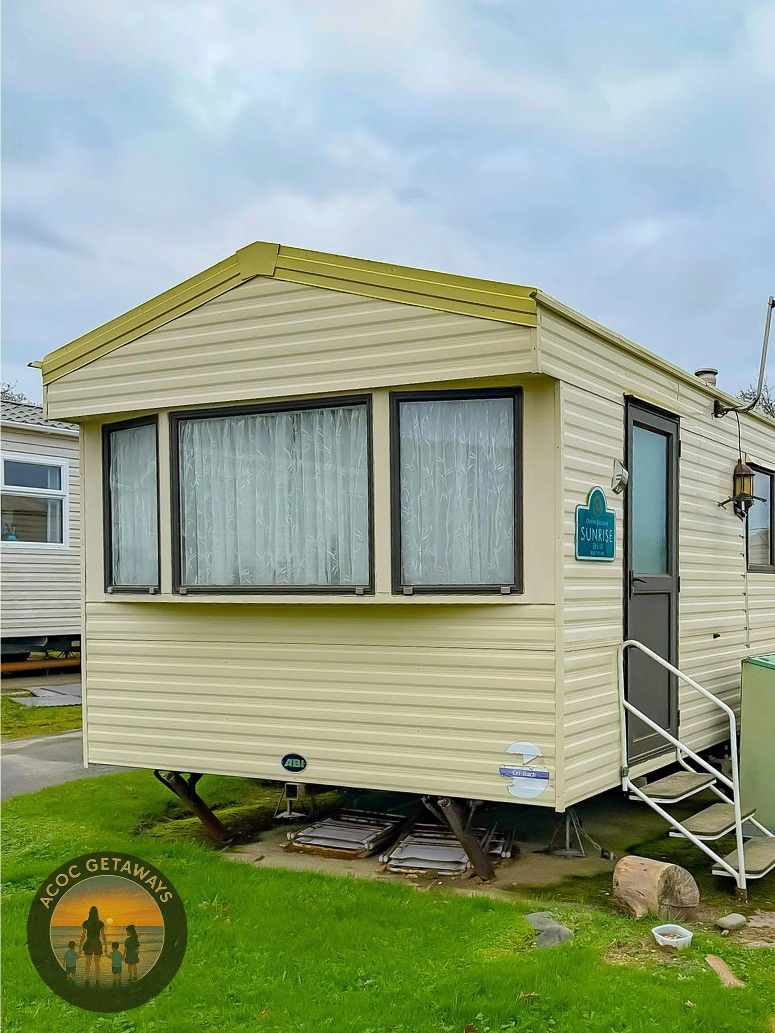 A beige mobile home with three large front windows, a dark door with a small set of stairs, and a sign that says 'Sunrise' on the side. The mobile home is elevated off the ground on metal supports with some stacked items underneath. The sky is cloudy, and the grassy area around it has rocks and a log. There is a logo in the bottom left corner that says 'ACOC GETAWAYS' with a silhouette of a family at sunset.