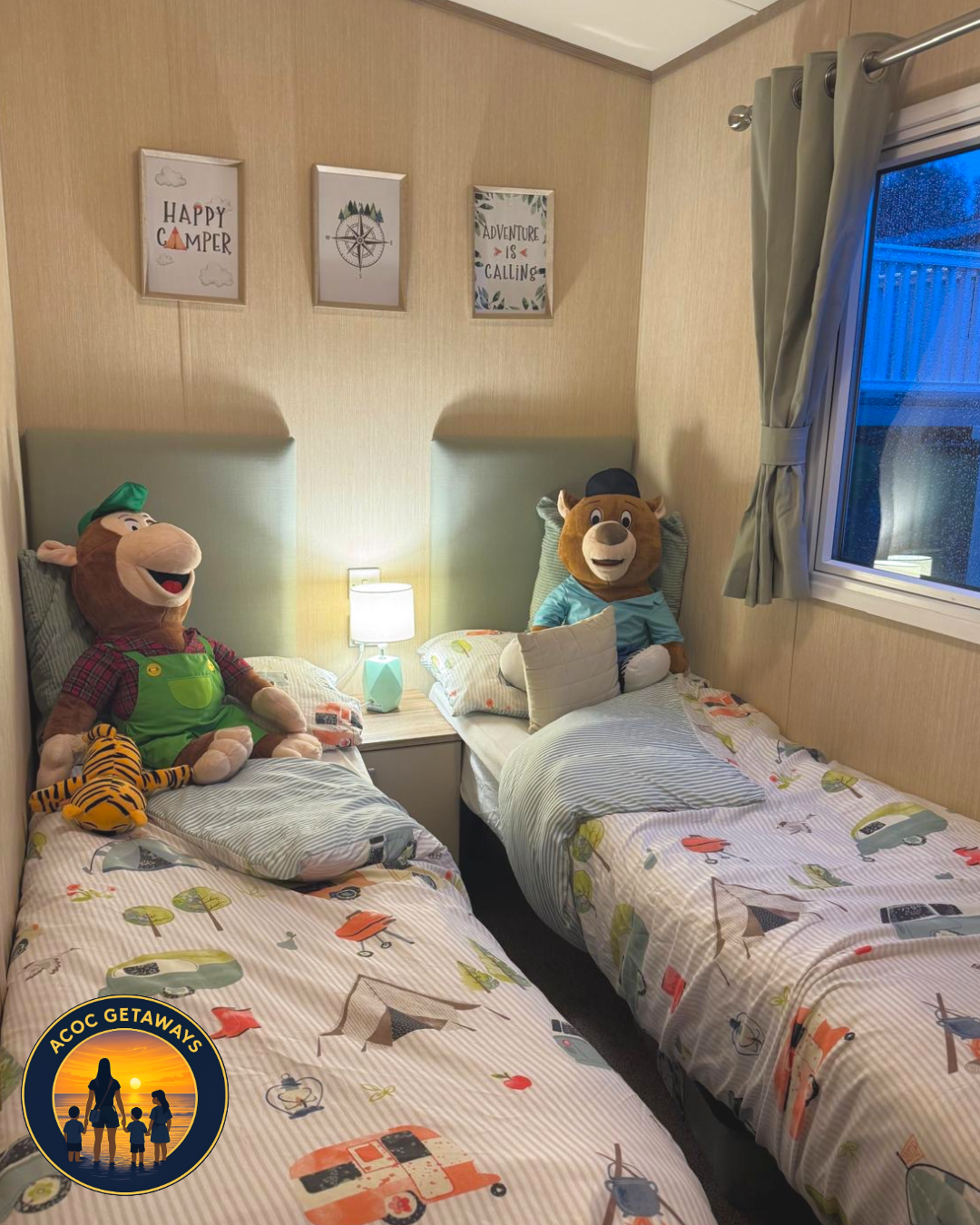 Two twin beds in a cozy room, each with colorful camping-themed bedding and plush toys of cartoon characters, with framed wall art above and a window with gray curtains to the side.