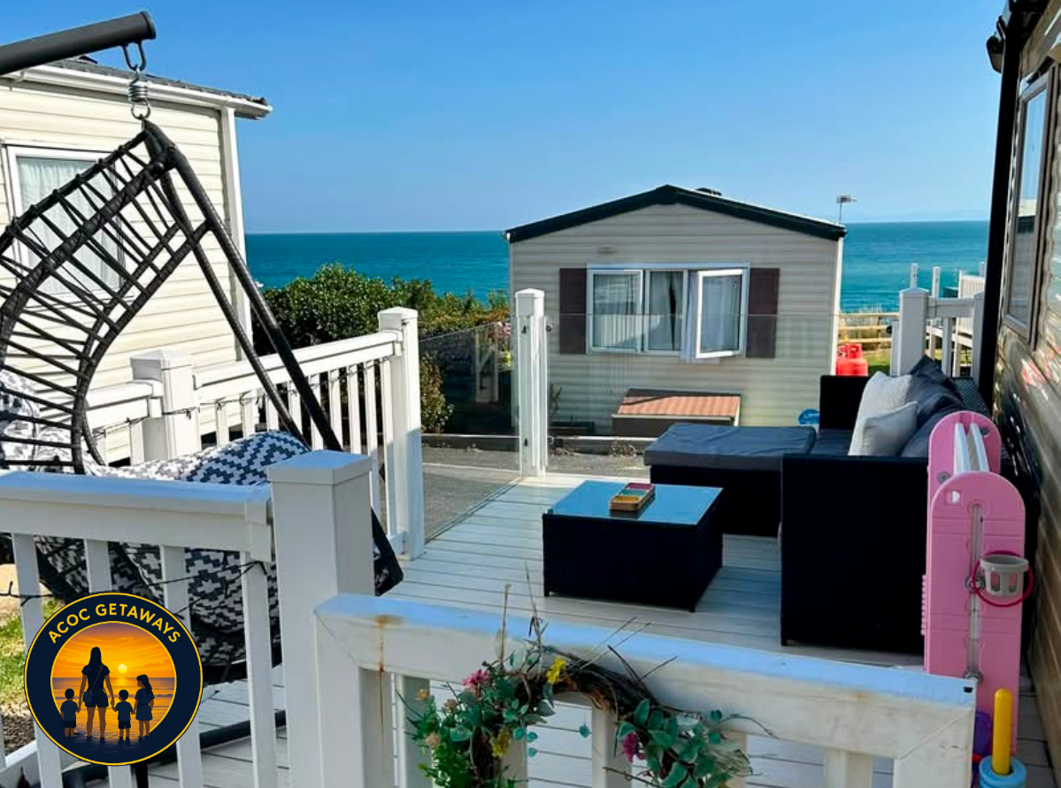 A balcony overlooking the ocean with outdoor furniture, a hanging egg chair, and a pink children's slide, against a bright blue sky.