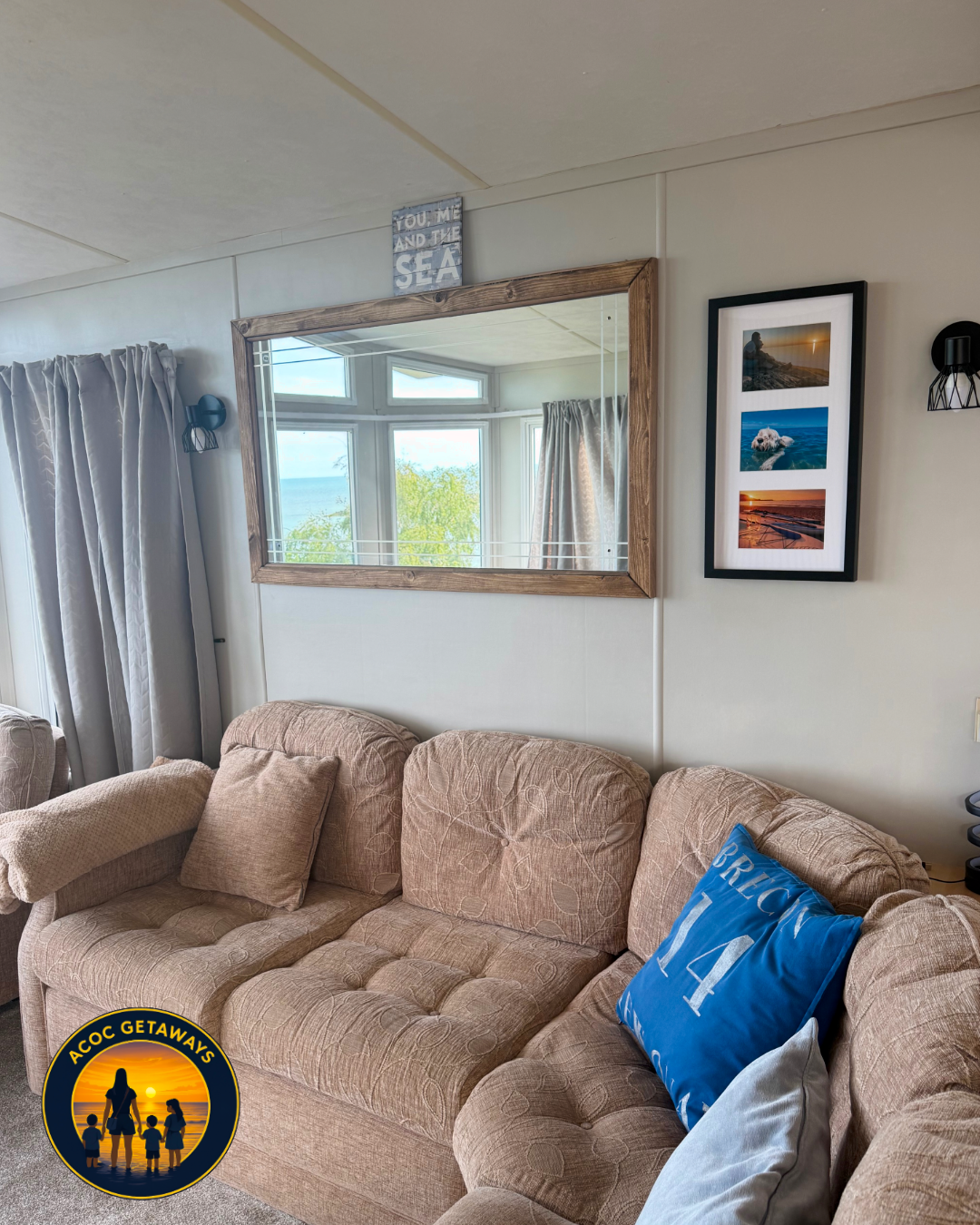 Living room with beige couch, blue pillow, mirror reflecting a window with a view of trees and the sea, framed group of beach photos, and a small sign that says ''You, Me, and the Sea''.