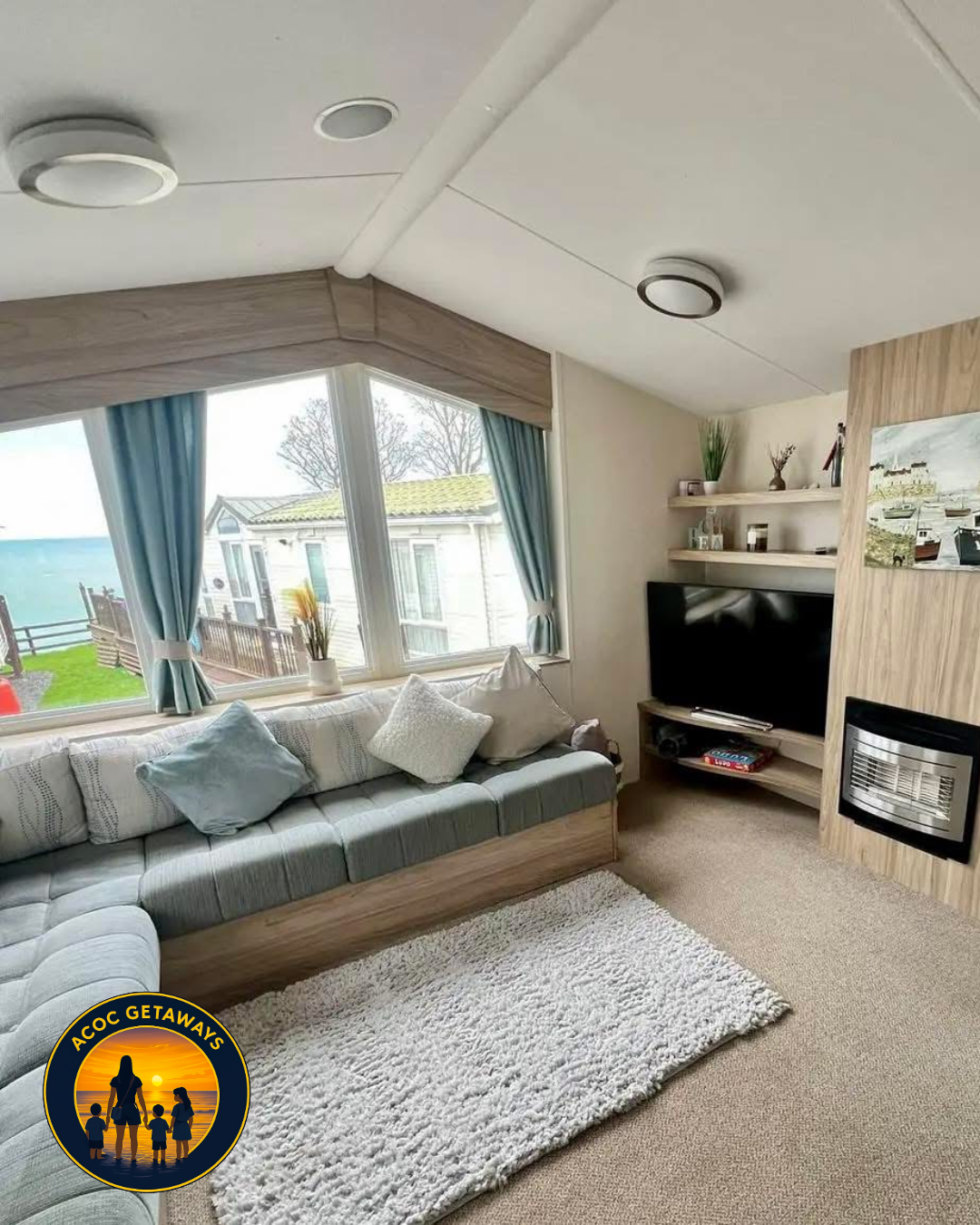 Cozy living room with large window view of the beach, light-colored sofa with pillows, fluffy rug, and television with shelving and decorative items.
