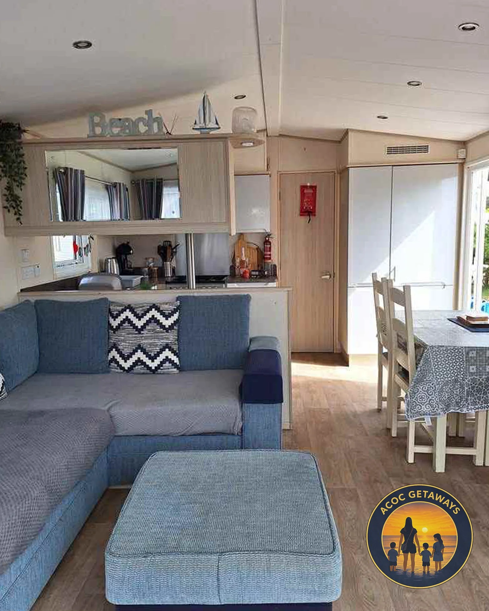 Living room and dining area in a vacation rental with a blue sectional sofa, a mix of cushions, wooden flooring, a dining table with chairs covered by a patterned tablecloth, and a kitchen with coffee maker and utensils. A sign spelling 'Beach' is on