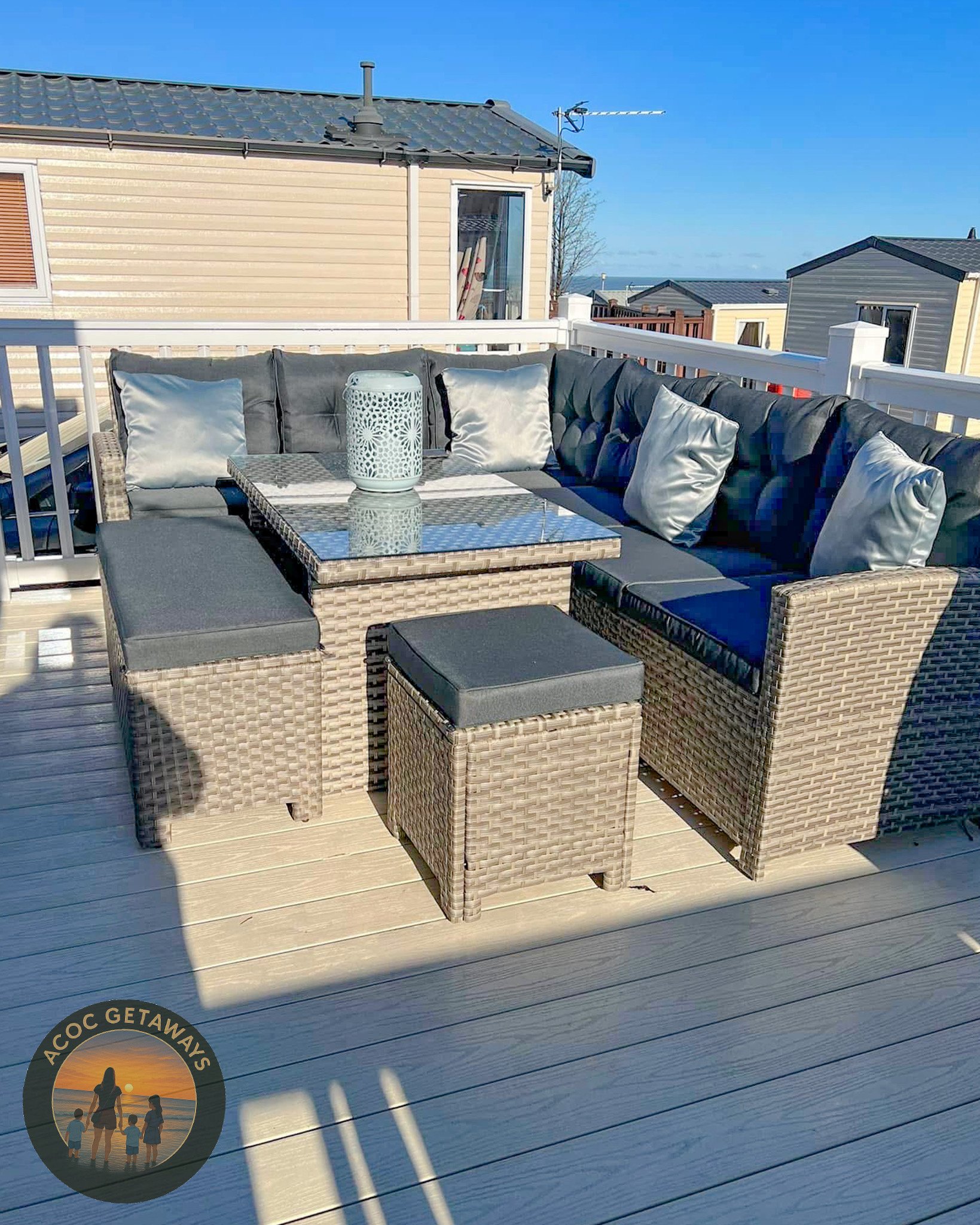 Outdoor patio with a wicker sectional sofa, grey cushions, and silver pillows, a glass-top table with a woven base, and a wicker ottoman, on a wooden deck with a white railing, under a clear blue sky.