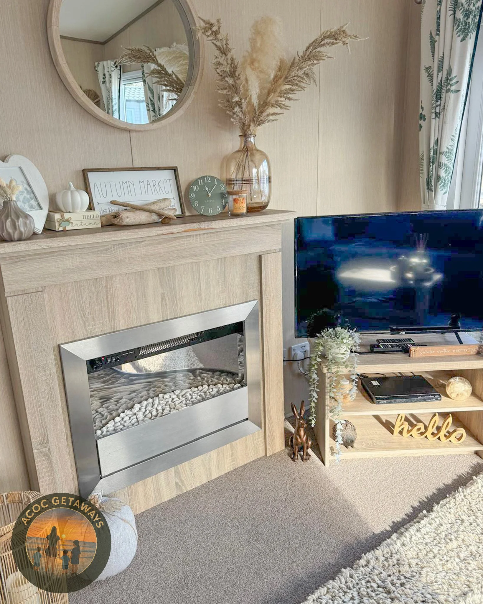 Living room with a fireplace, TV, and autumn-themed decor including dried pampas grass in a glass vase, a pumpkin, a framed sign reading 'Autumn Market', a round clock, and fall-themed ornaments on the mantel. There is a small plush pumpkin and a woo