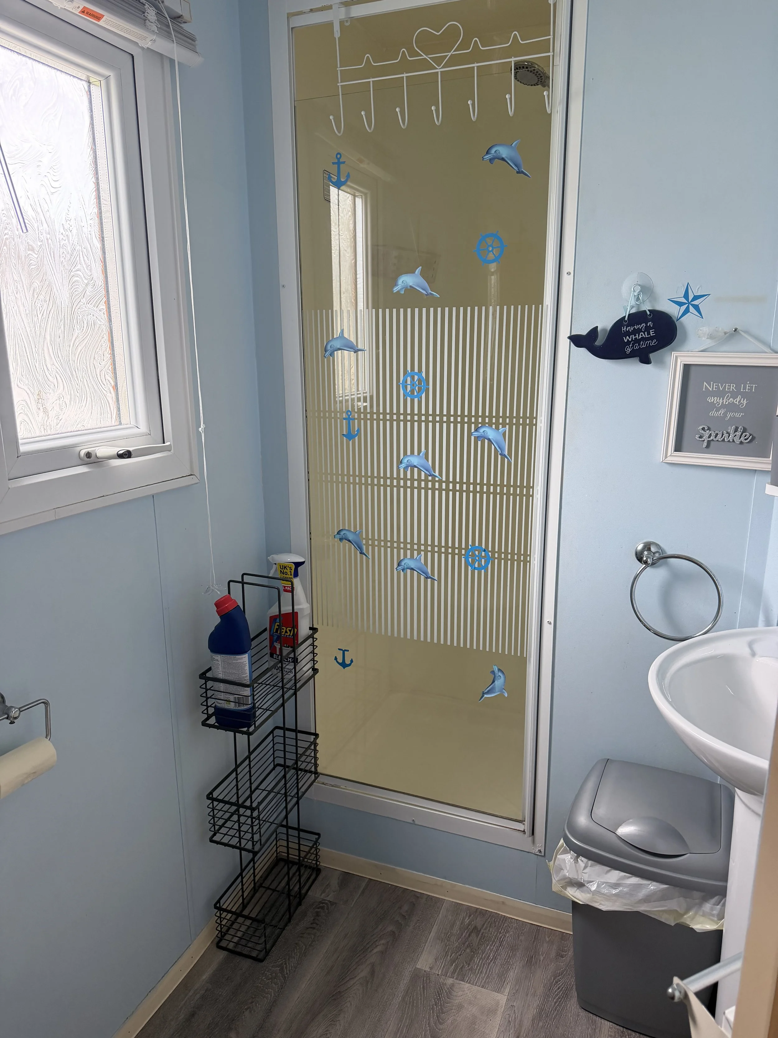 A bathroom with a light blue wall, a window, a shower with a sea-themed sticker curtain, a black wire storage rack, a trash bin with a trash bag, a white sink, and decorative signs about whales and sparking.