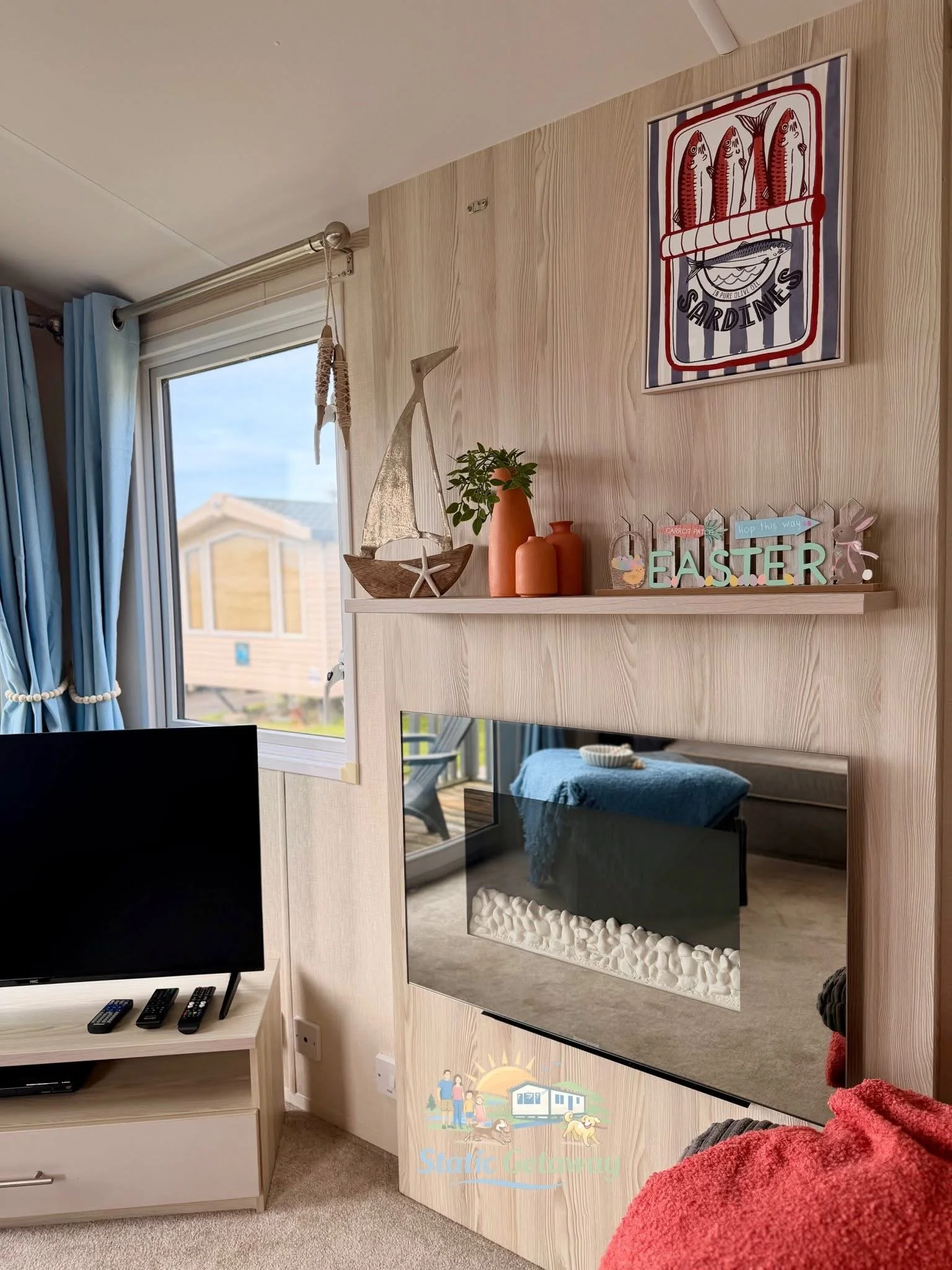 A modern living room with a wall-mounted TV, decorative shelves with sea-themed decor, a window with blue curtains, and a fireplace below the mirror. There is a red blanket or towel in the foreground.