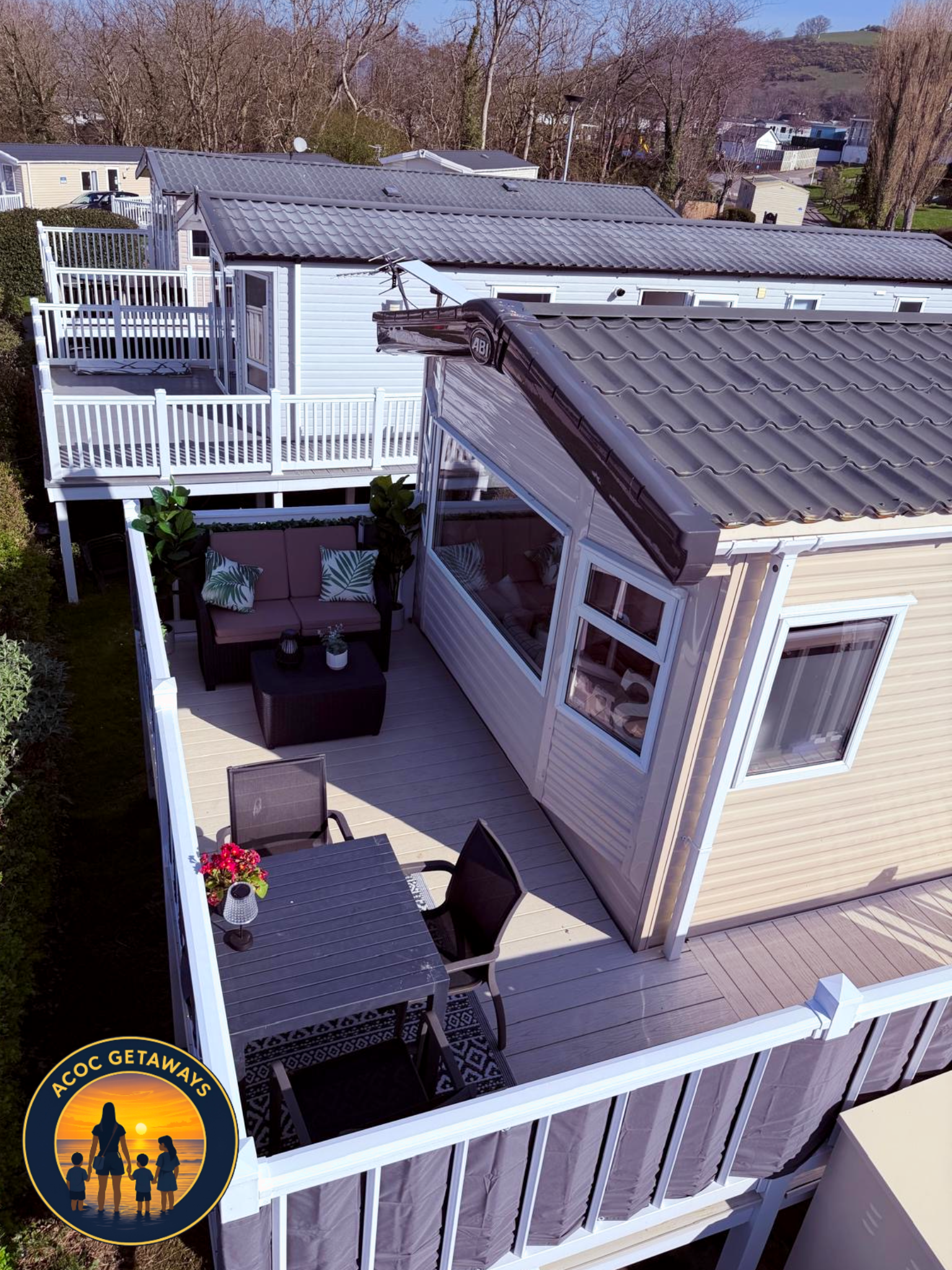 A view of a white house with a deck, outdoor furniture, potted plants, and a railing, set against a backdrop of trees and neighboring houses.