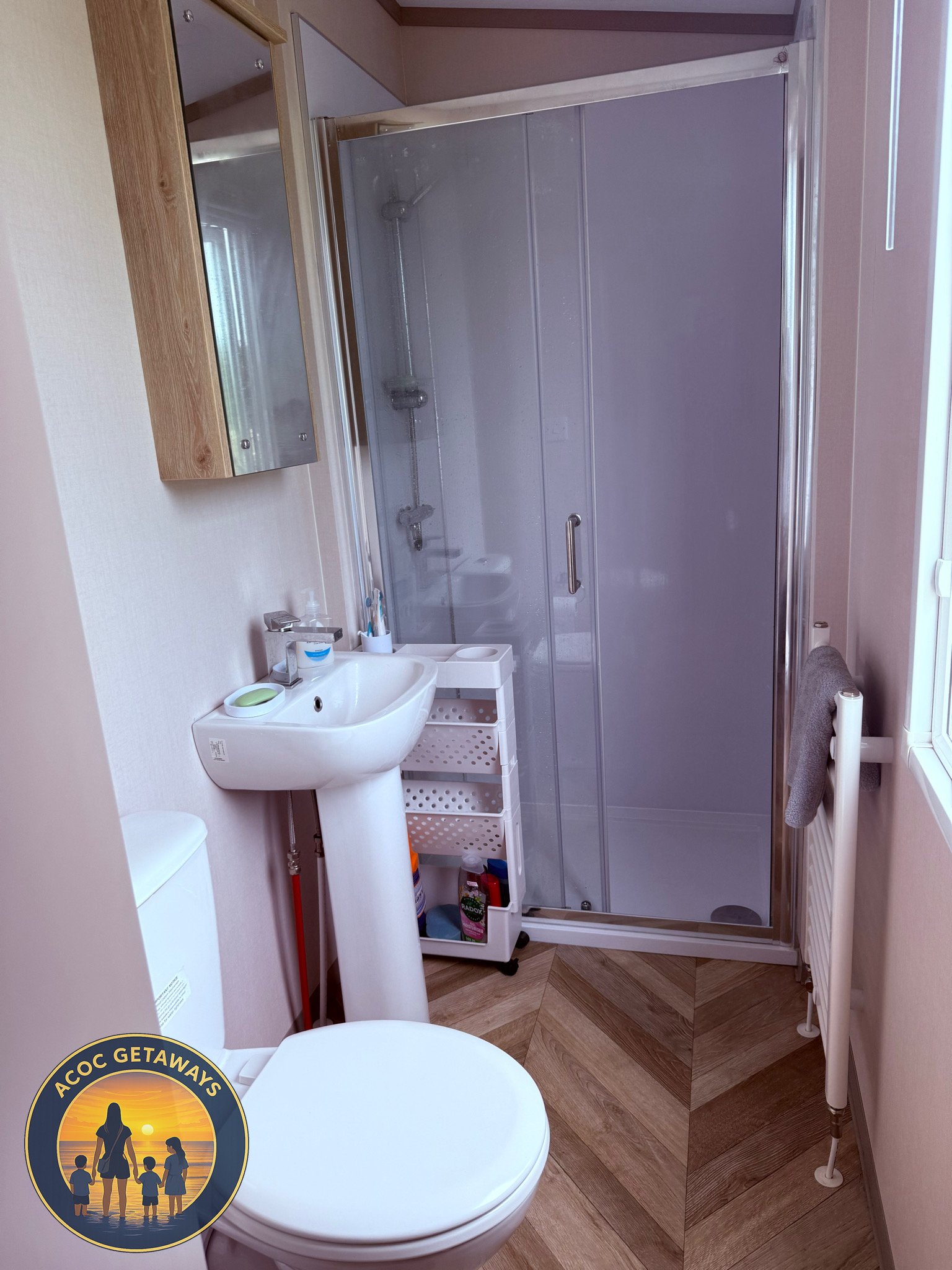 Small bathroom with a shower stall with glass door, a white pedestal sink with storage underneath, a mirror cabinet above, a toilet, and a window. There are personal care items on the sink and a towel hanging on a radiator.