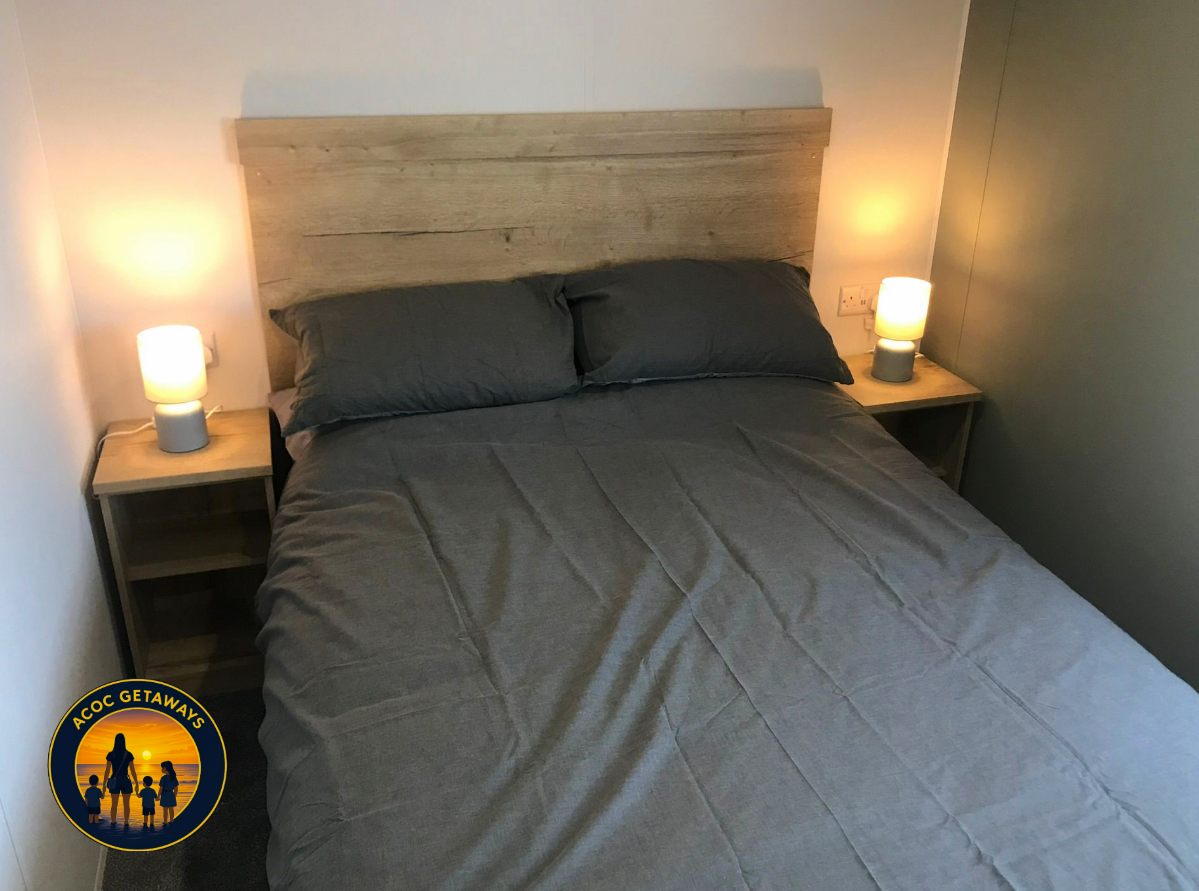 A neatly made bed with dark gray sheets and pillows in a bedroom with two nightstands, each with a lit table lamp. The headboard is made of light wood, and the walls are painted white and gray.