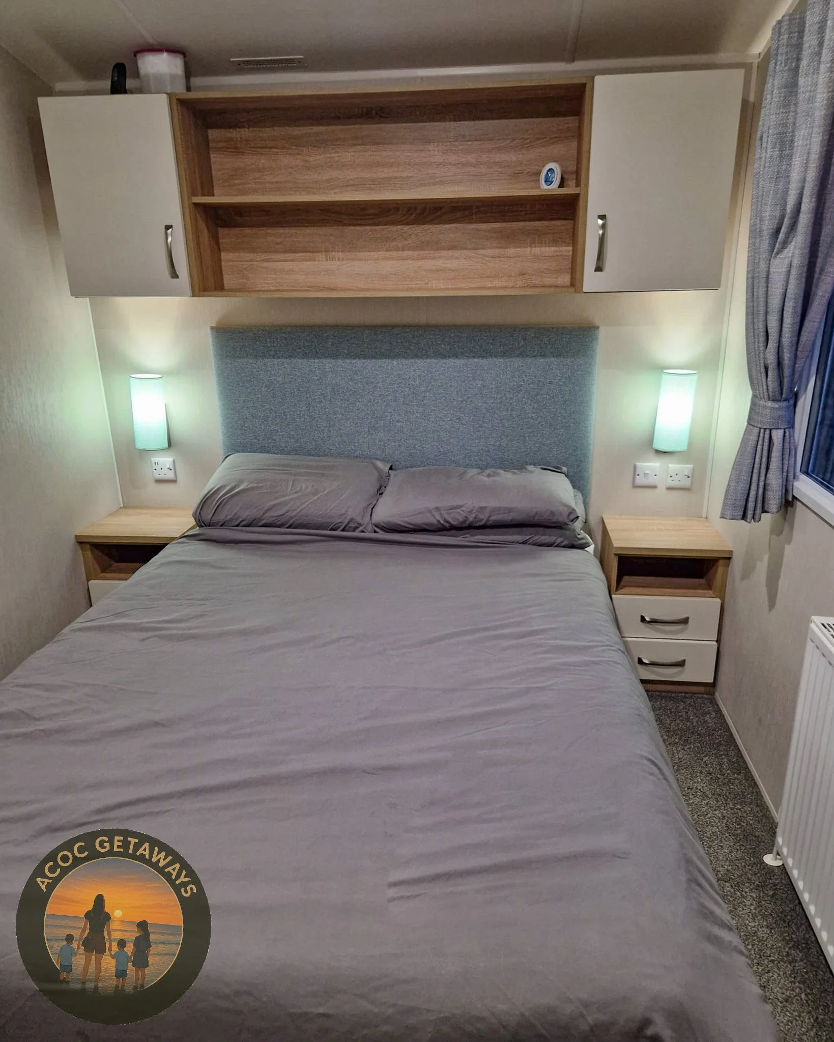 A neatly made bed with gray sheets and pillows, blue upholstered headboard, two wooden nightstands with drawers, wall-mounted lamps, a window with blue curtains, and a wooden overhead shelf with a few items on top.