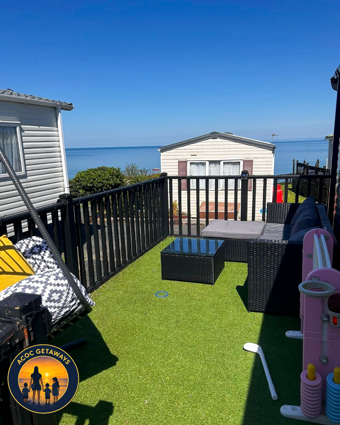 A colorful outdoor balcony with green artificial grass flooring, black wicker furniture including a sofa and small table, overlooking a scenic view of a blue ocean and sky with neighboring beach houses, some children's toys, and a logo in the bottom 