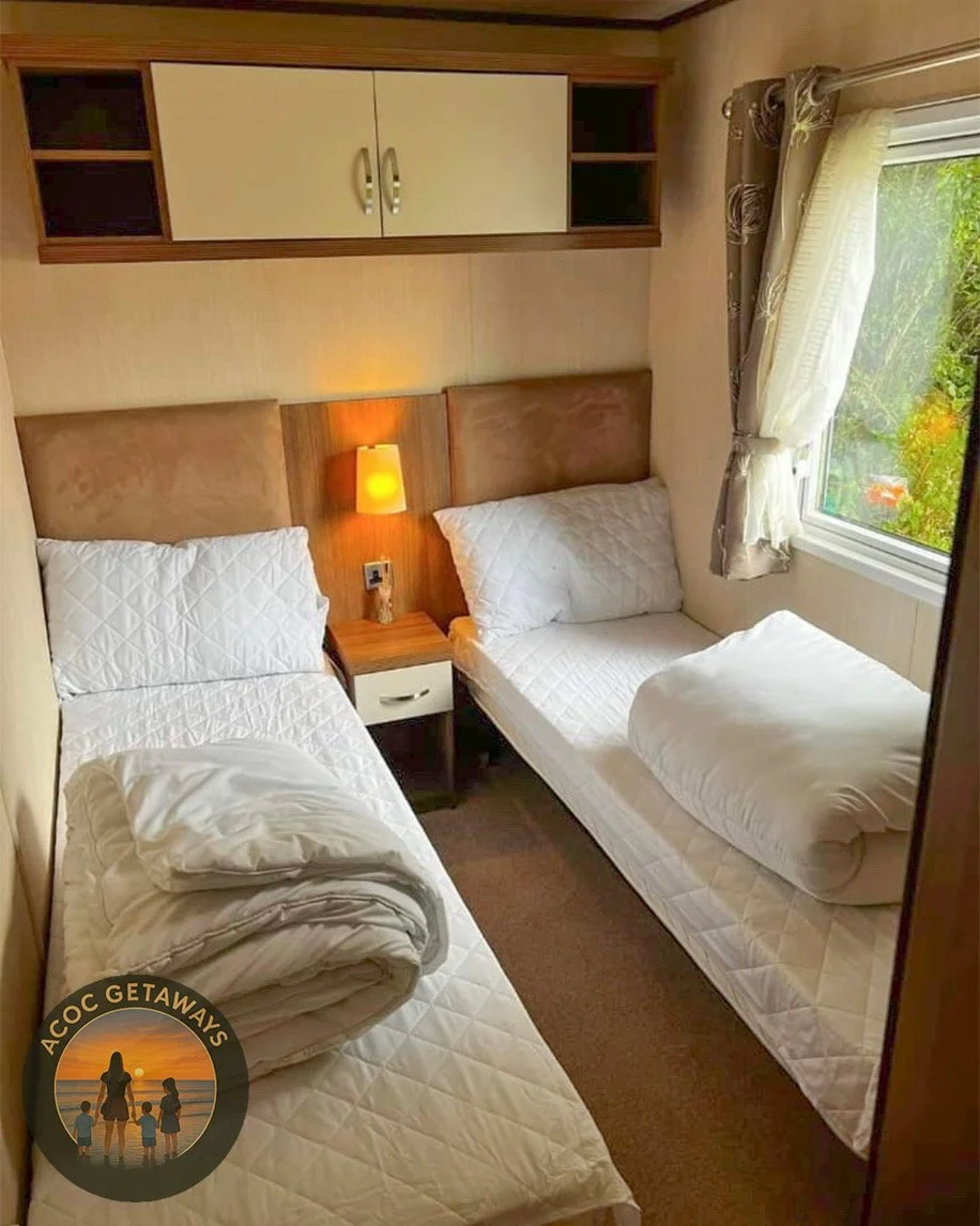 A small bedroom with two single beds with white quilted bedding and pillows. A window with curtains shows greenery outside. A wood-paneled headboard runs behind both beds, with a nightstand and lamp between them. Overhead, there are rectangular storage cabinets with beige doors and open shelves.