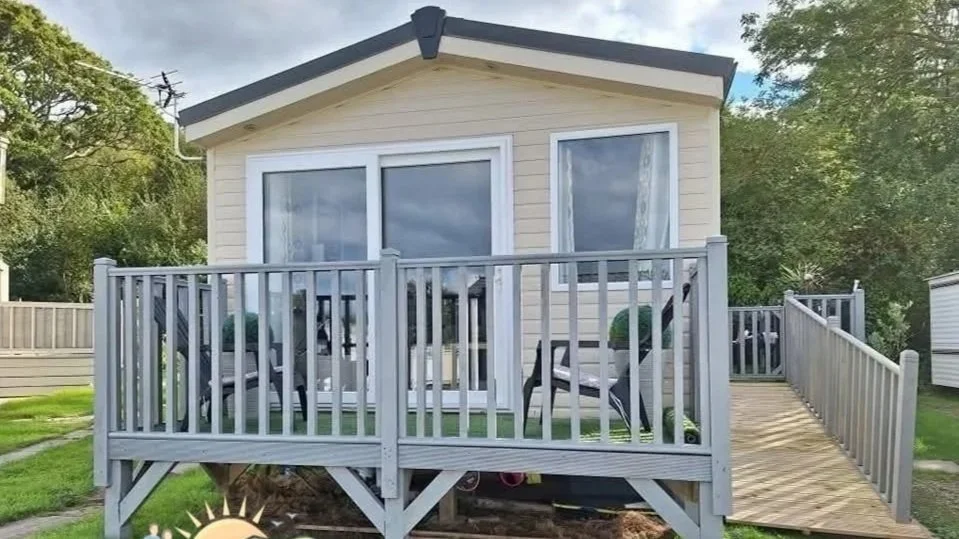 Small beige house with a wooden deck and gray railing, featuring large sliding glass doors and one window, surrounded by green trees and grass.