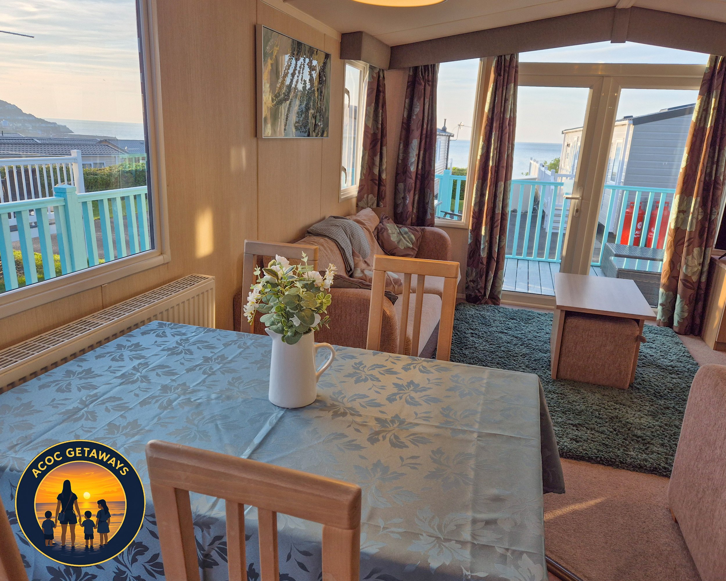 Living room with sliding glass door leading to a balcony overlooking the ocean, floral curtains, a pink couch, a coffee table, and a rug. A dining table with a floral tablecloth and a vase of flowers is in the foreground, with a window showing a coas