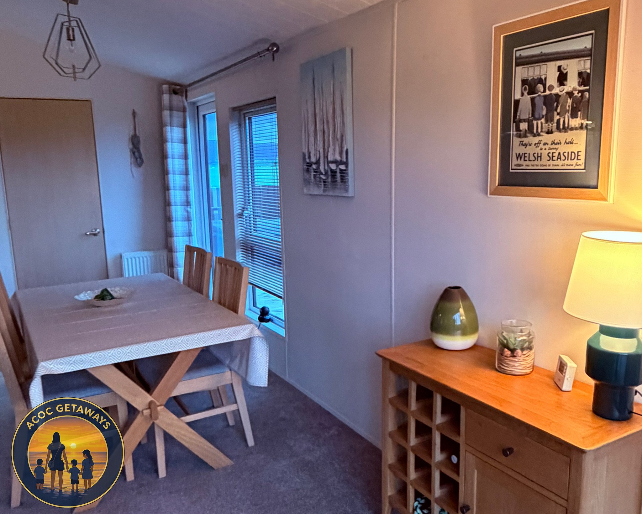 A cozy living room with a dining table covered with a beige tablecloth, surrounded by four wooden chairs. There is a door on the left, a large window with blinds and patterned curtains, and framed pictures on the wall. A wooden sideboard with a decor