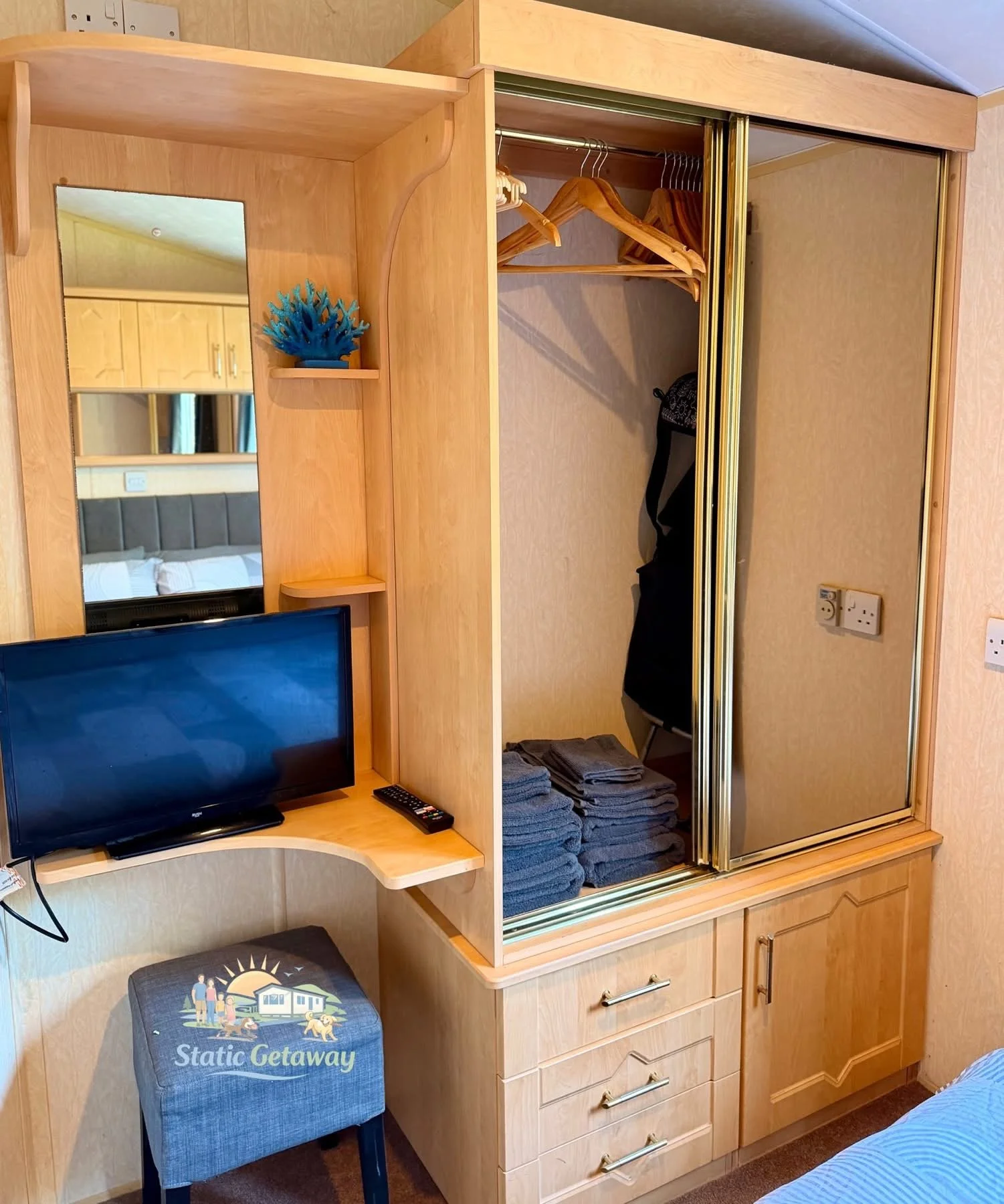 Hotel room with wooden furniture, including a wardrobe with mirrored sliding doors, a small desk with a TV, and folded towels. A blue decorative plant is on a small shelf, and a blue upholstered chair with a logo that says 'Static Getaway' is nearby.