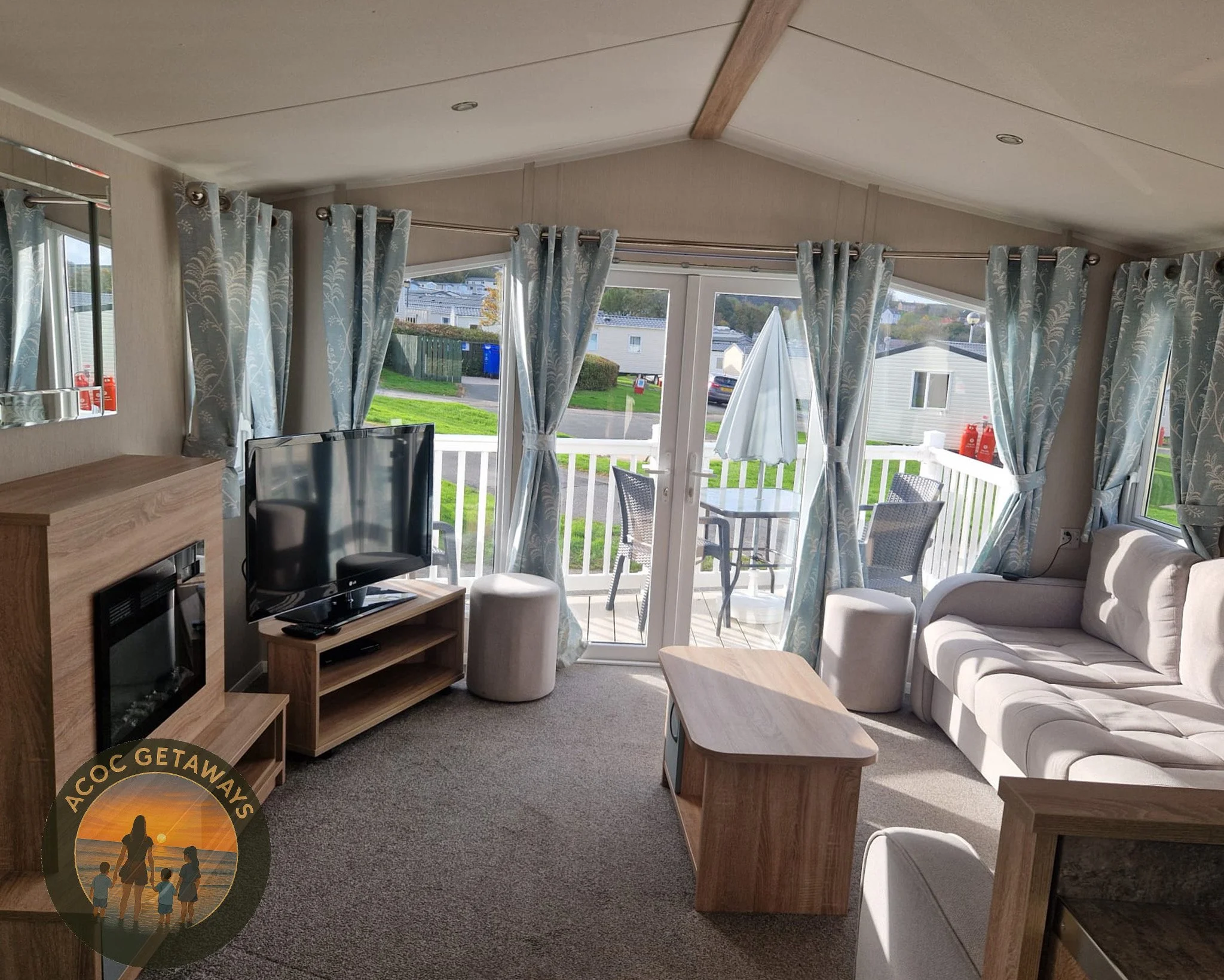 Living room with large windows and glass door leading to balcony with outdoor table and chairs, beige sofa, TV, and fireplace, with light blue patterned curtains.