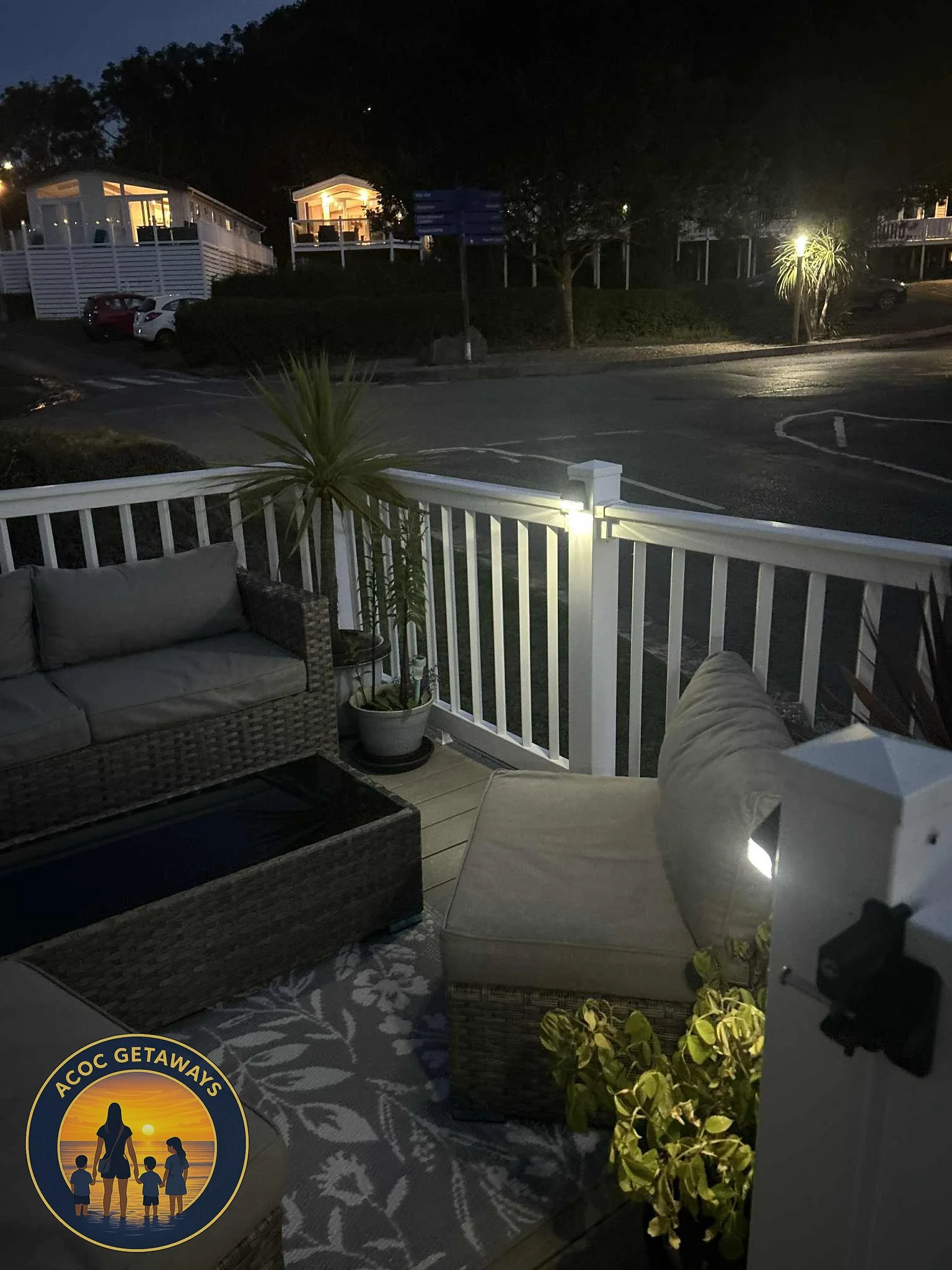 A nighttime view from a porch with outdoor furniture, including a woven sofa, an ottoman, and a potted plant, overlooking a street with houses and parked cars.