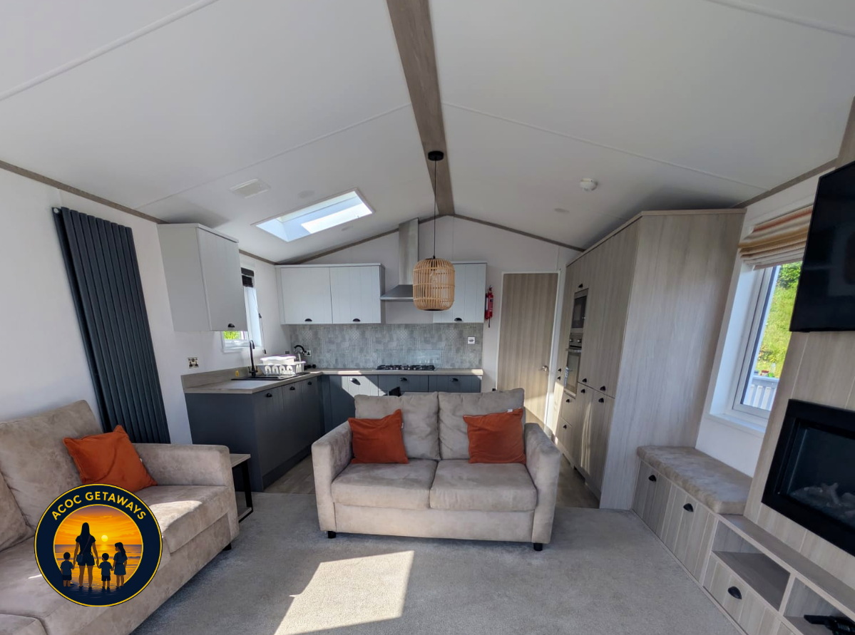 Living room and kitchen area in a modern tiny home with beige sofas, orange cushions, a built-in window seat, and a kitchen with white and gray cabinetry, a skylight, and a hanging wicker light fixture.